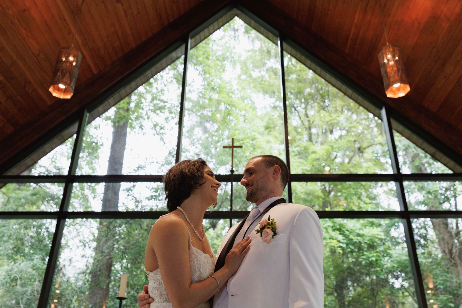 Bride and groom share romantic moment inside chapel with large triangular stained glass forest window.