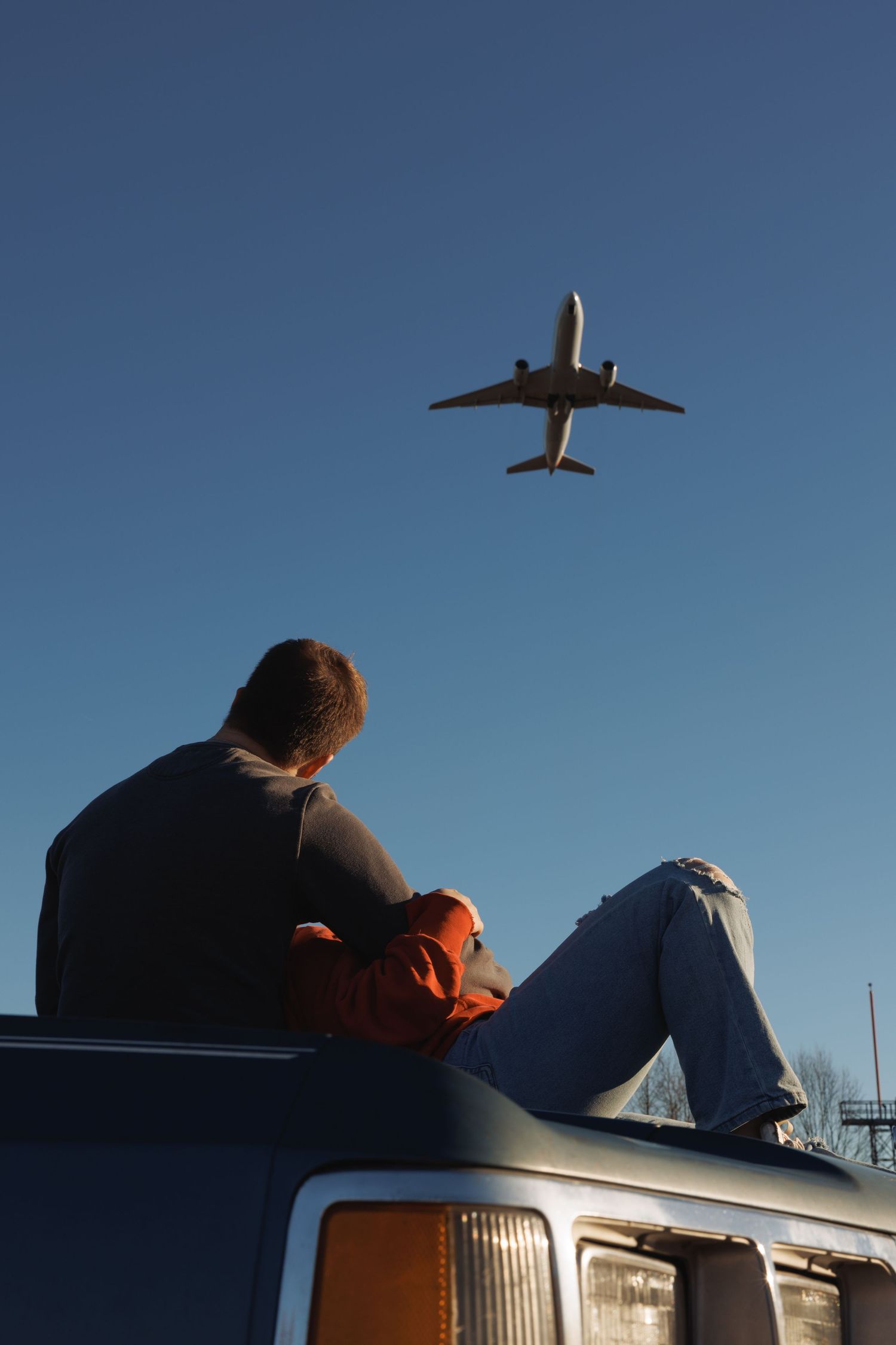 Man sitting on top of a van watching a commercial airplane flying overhead against a clear blue sky.
