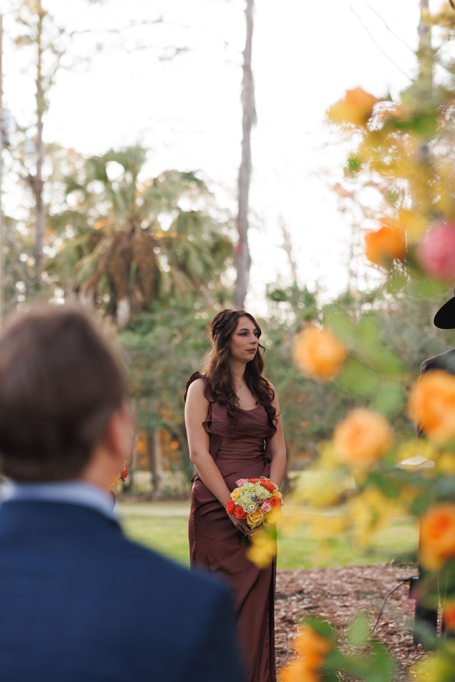 Bridesmaid in brown gown holds colorful bouquet surrounded by orange roses at outdoor ceremony.