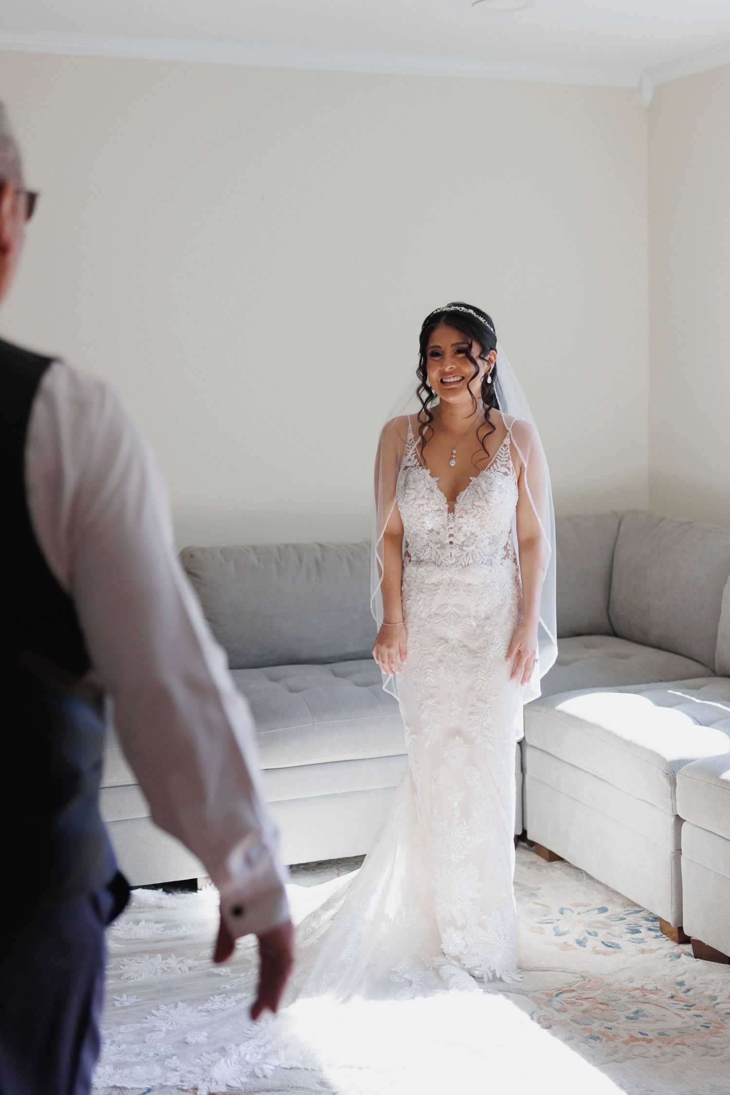 Smiling bride in lace wedding gown and veil stands in sunlit room as someone approaches her.