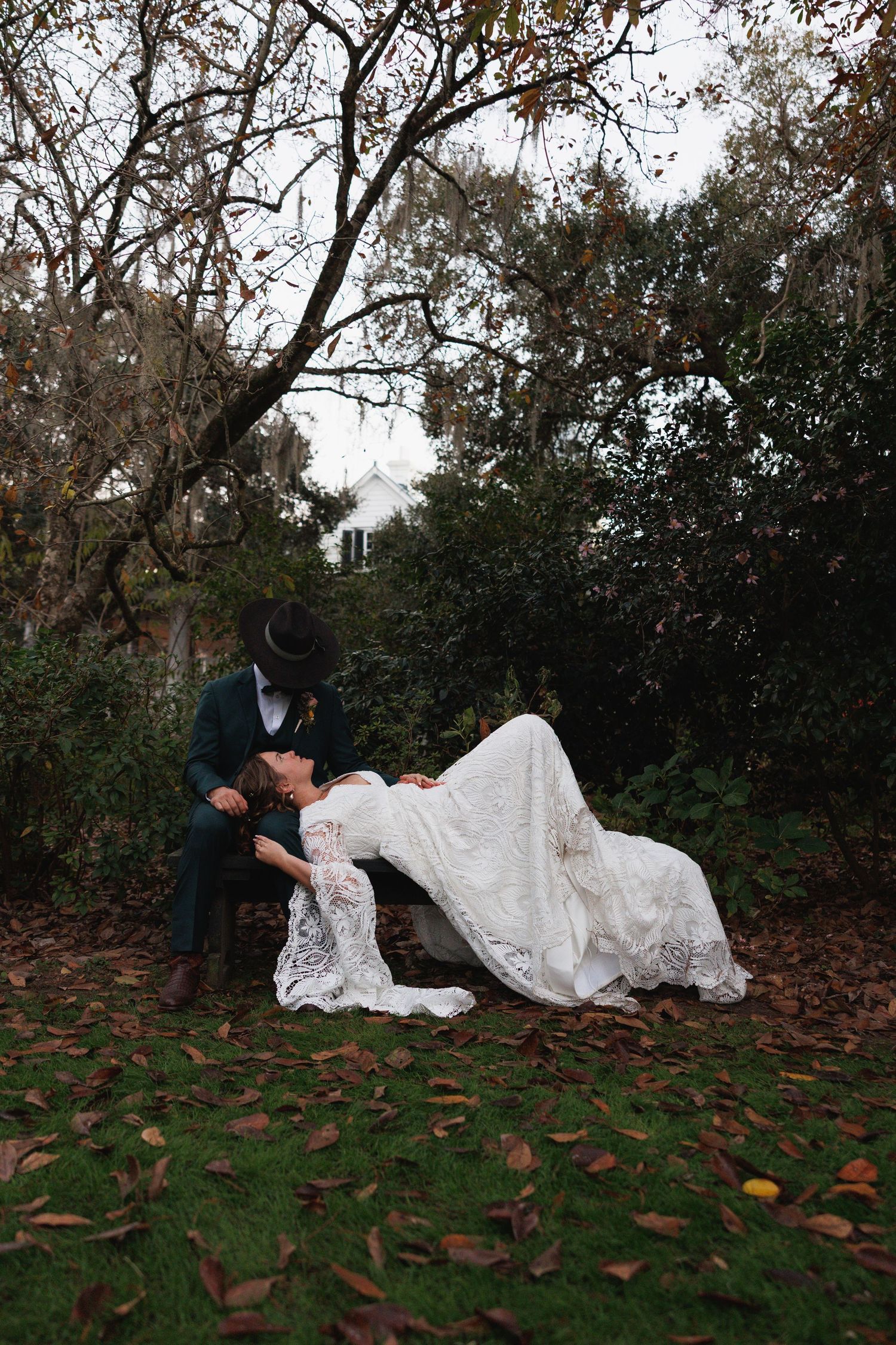 Bride and groom sharing romantic dip kiss on bench in autumn garden with fallen leaves.