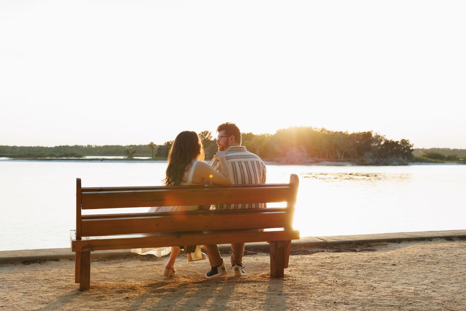 Couple sitting on a wooden bench watching a golden sunset over a calm lake with lush green trees in the background.