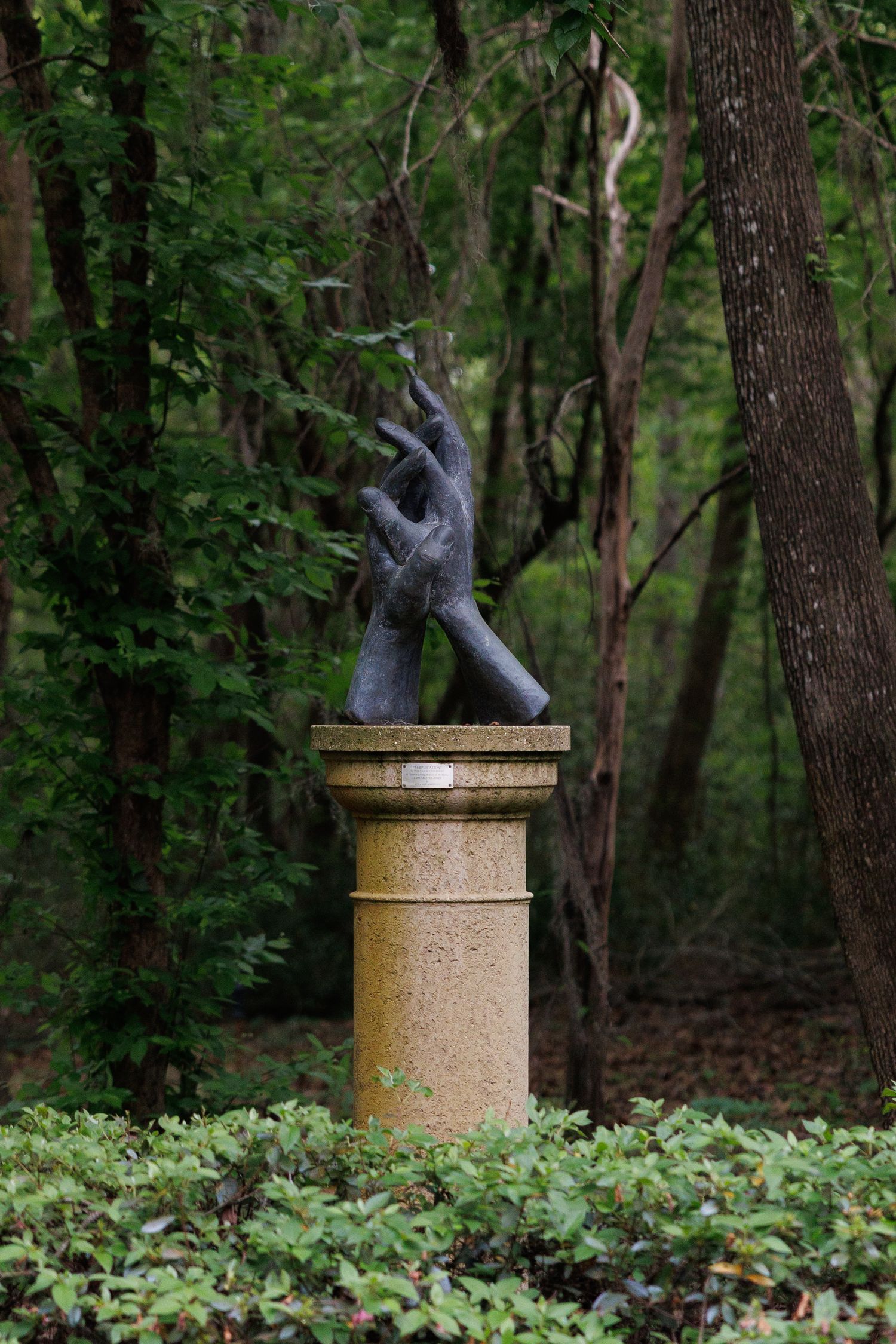Dark bronze sculpture of two hands on a stone pedestal surrounded by lush green forest trees.