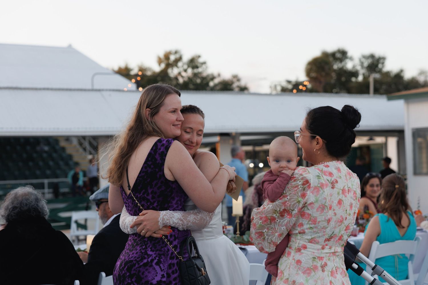 Wedding guests hugging and holding baby outdoors at evening reception venue.