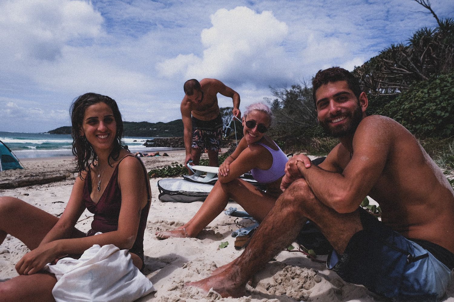 Group of friends relaxing on a tropical beach, smiling at camera with ocean waves and lush greenery in background.
