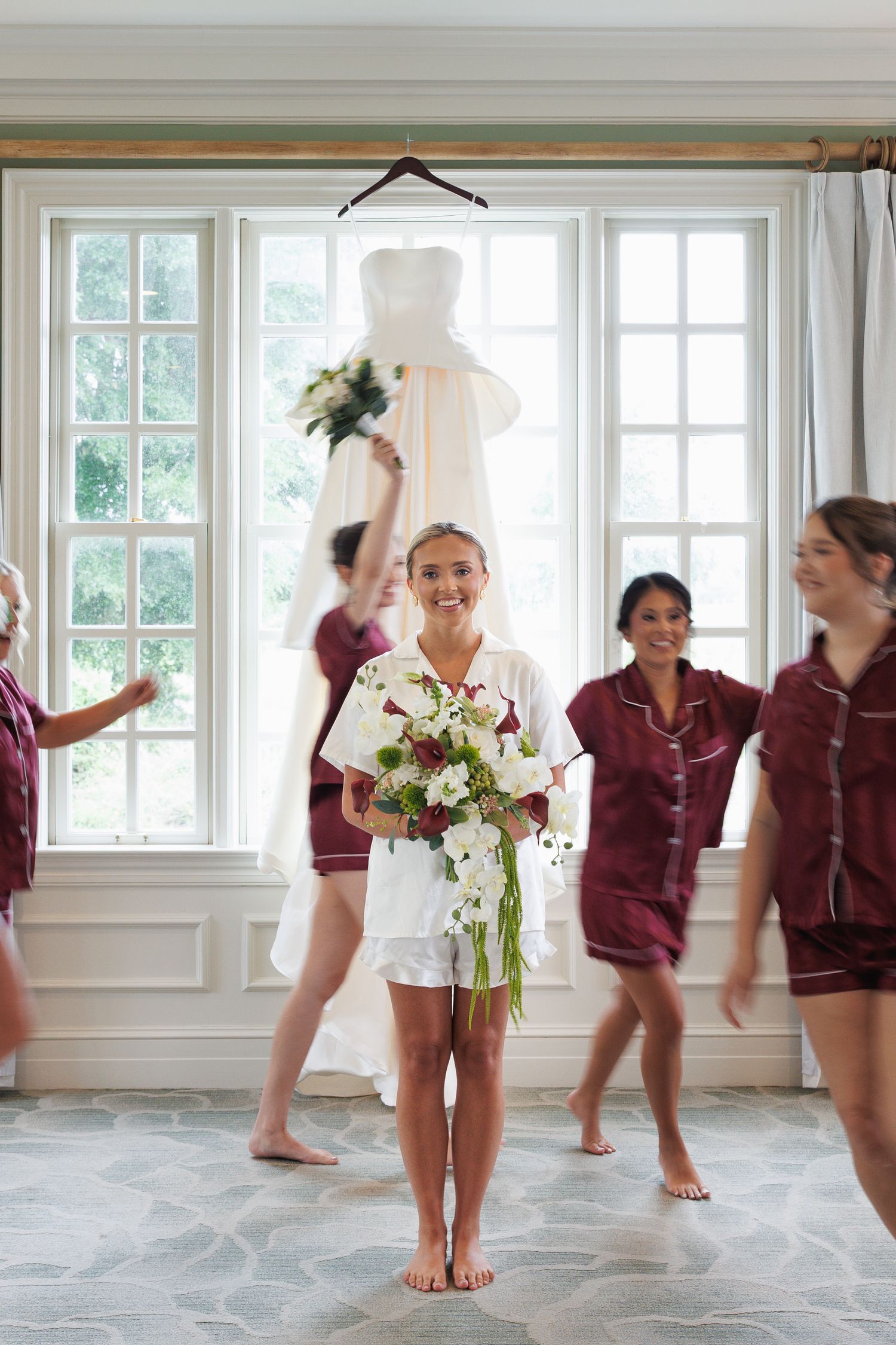 Bride in white pajamas holds bouquet surrounded by bridesmaids in burgundy sets before wedding.