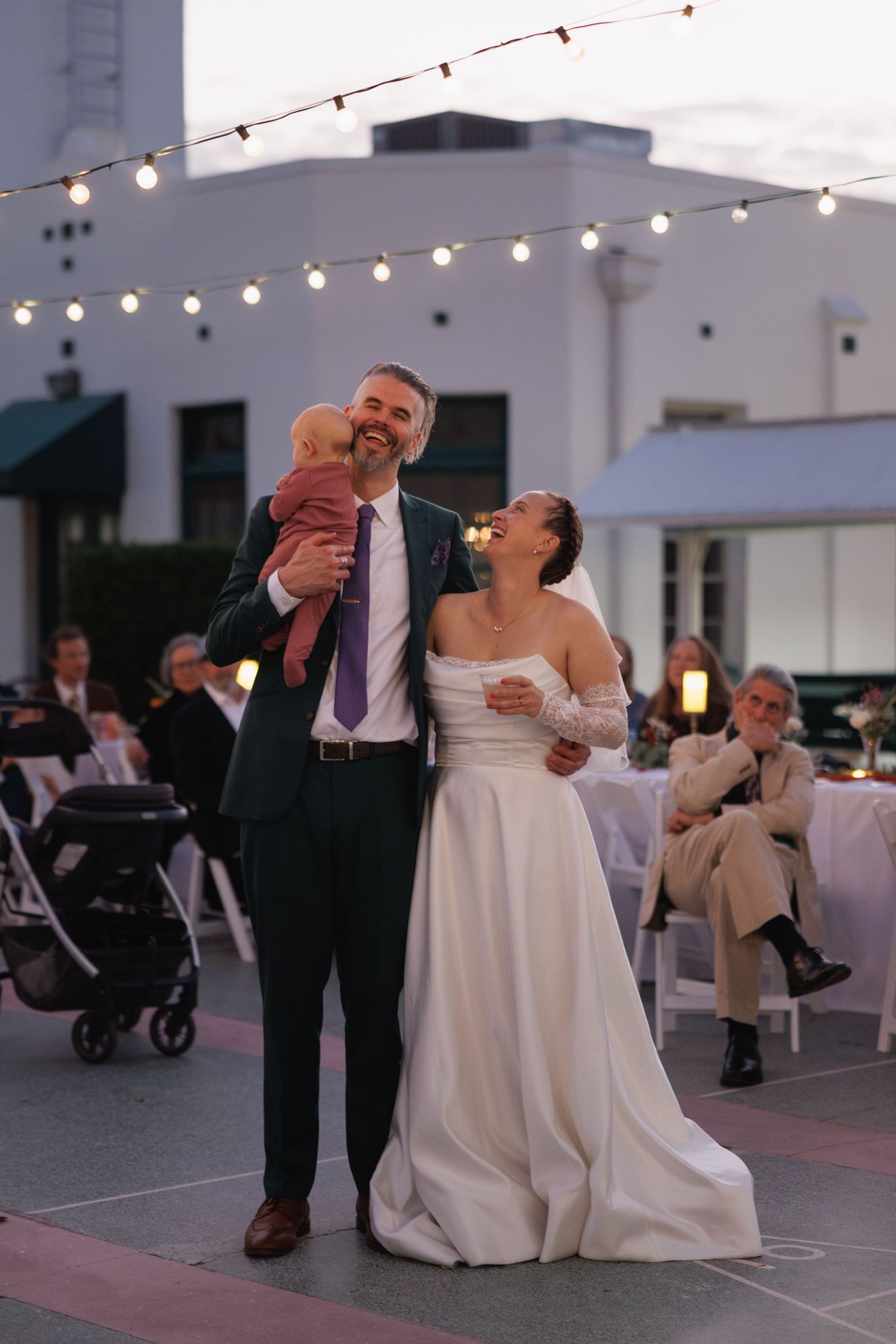 Smiling groom holding baby beside bride under string lights at outdoor wedding reception.