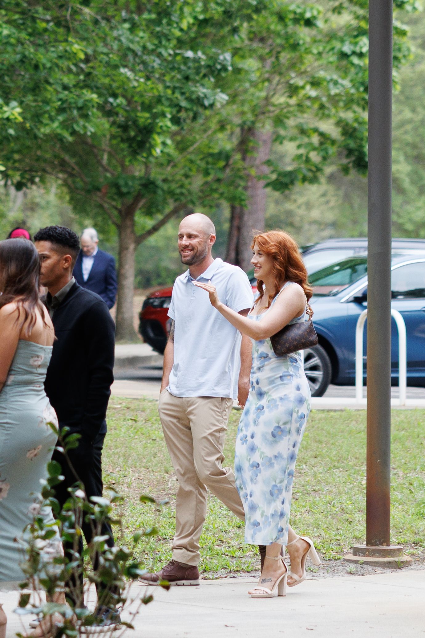 A bald man in a white shirt and khaki pants walks with a redhead woman in a floral dress outdoors.