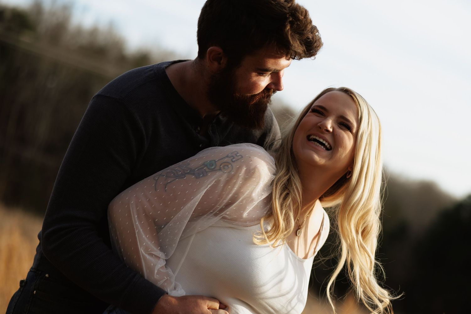 A bearded man embraces a laughing blonde woman in a white dress outdoors in a sunny field.