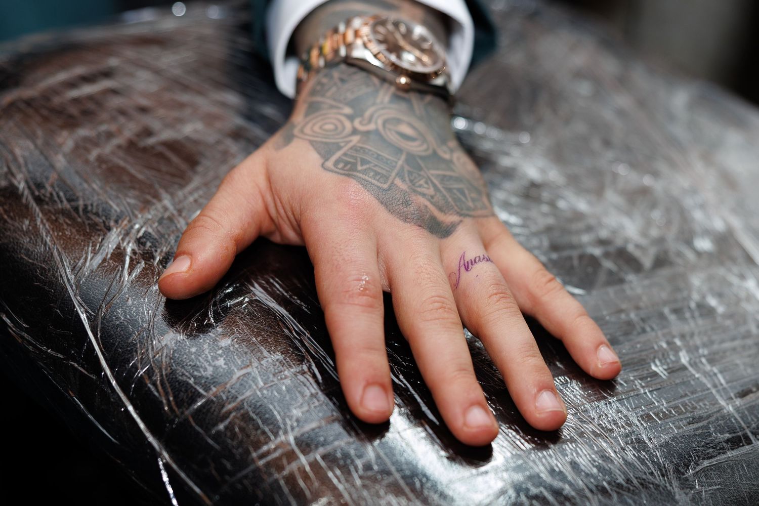 Close-up of tattooed hand with wedding ring resting on silver textured surface wearing gold watch.