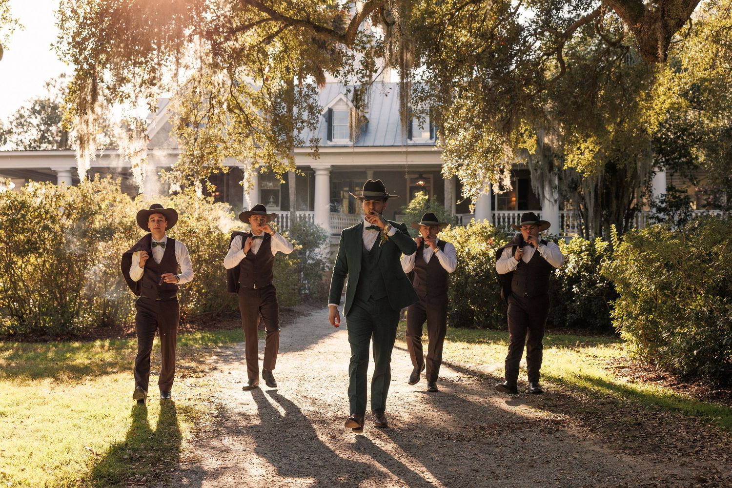 Groomsmen in dark vests and hats walking confidently down a sunlit path toward a historic Southern plantation home.