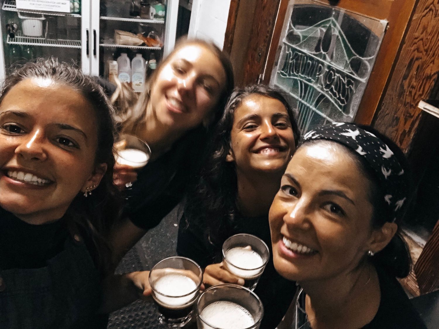 Four smiling women taking a selfie together while holding glasses of beer at a cozy indoor gathering.