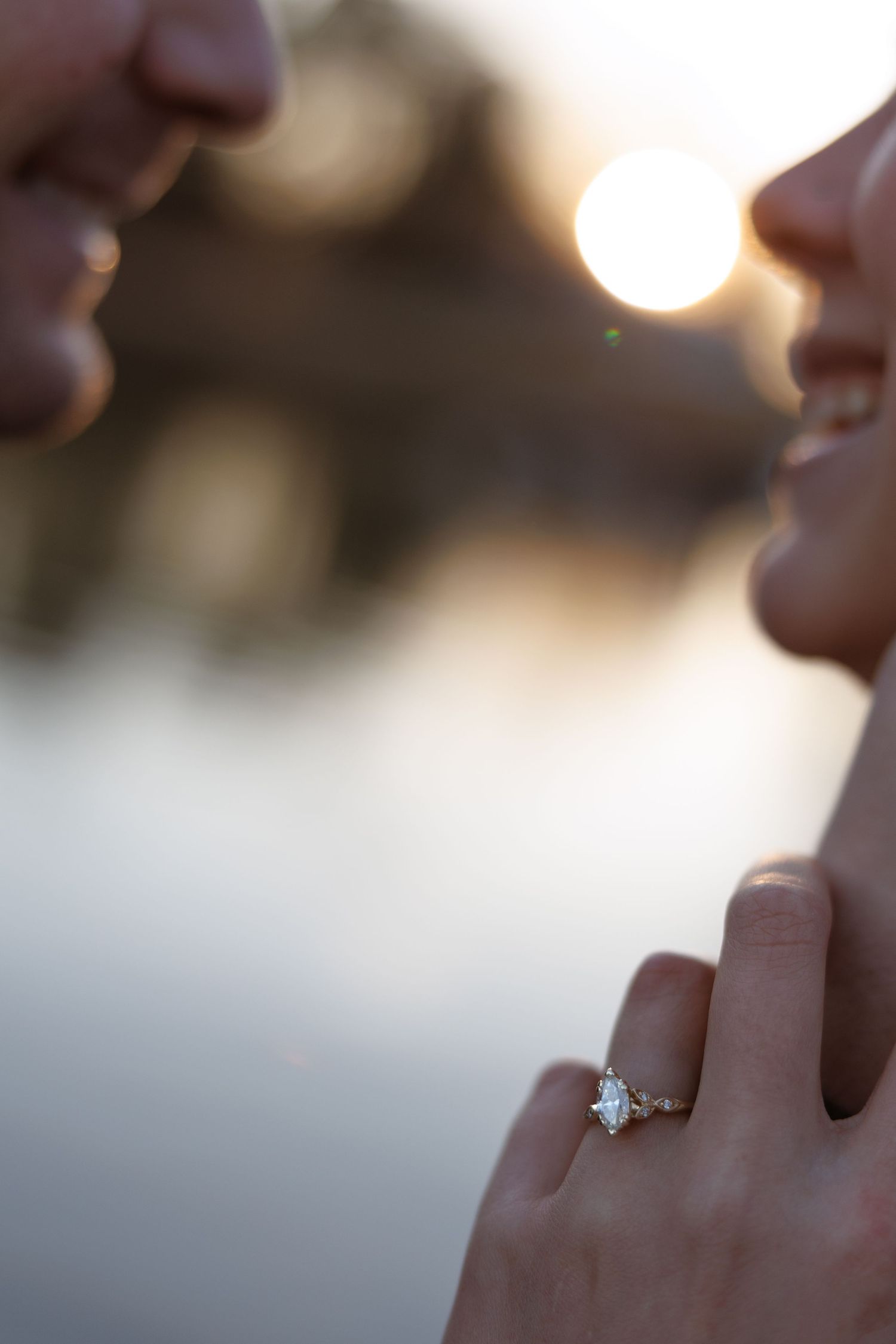 Close-up of an engagement ring on a woman's finger as a couple faces each other at sunset near water.