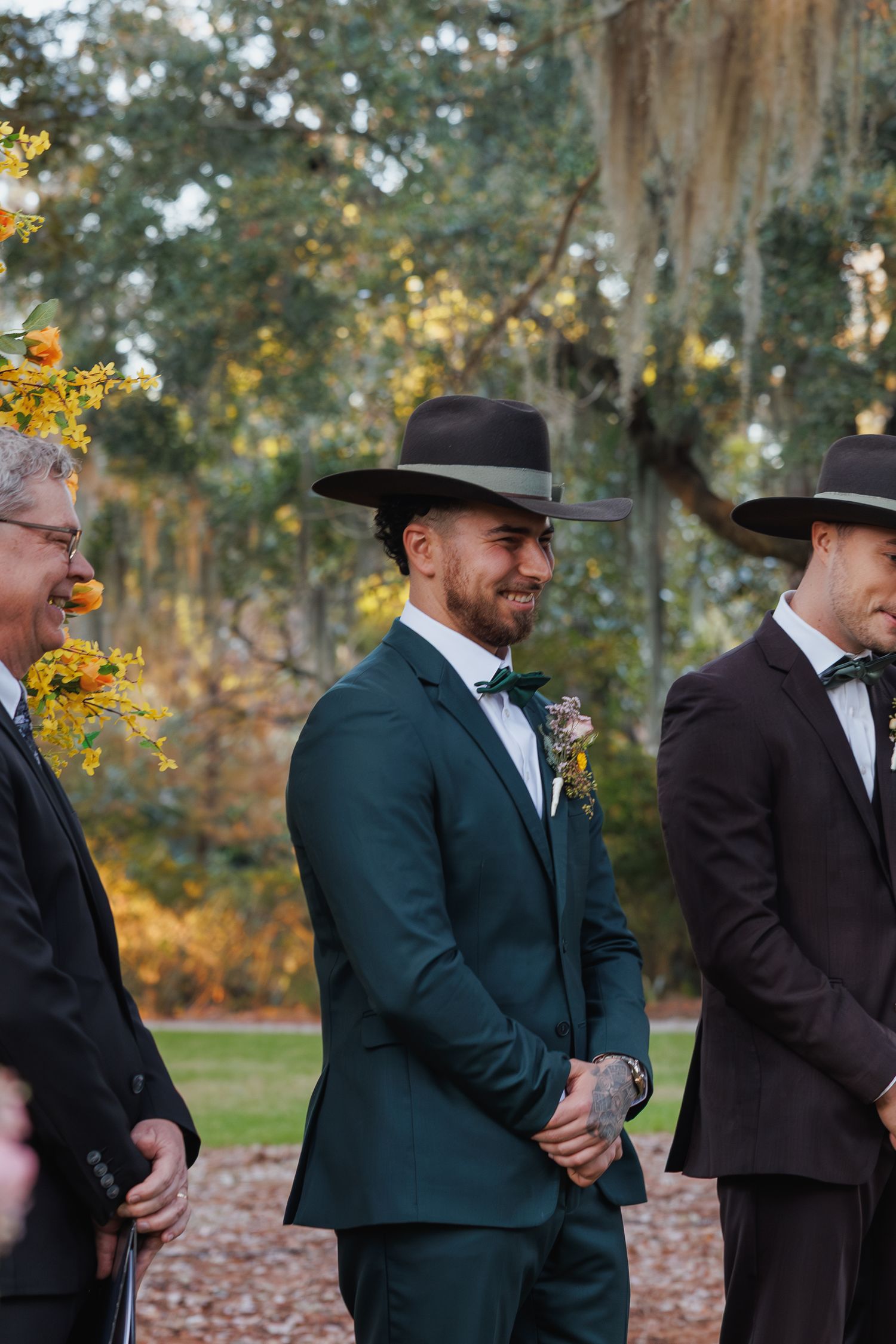 Groom in green suit and black cowboy hat smiling at outdoor wedding ceremony.