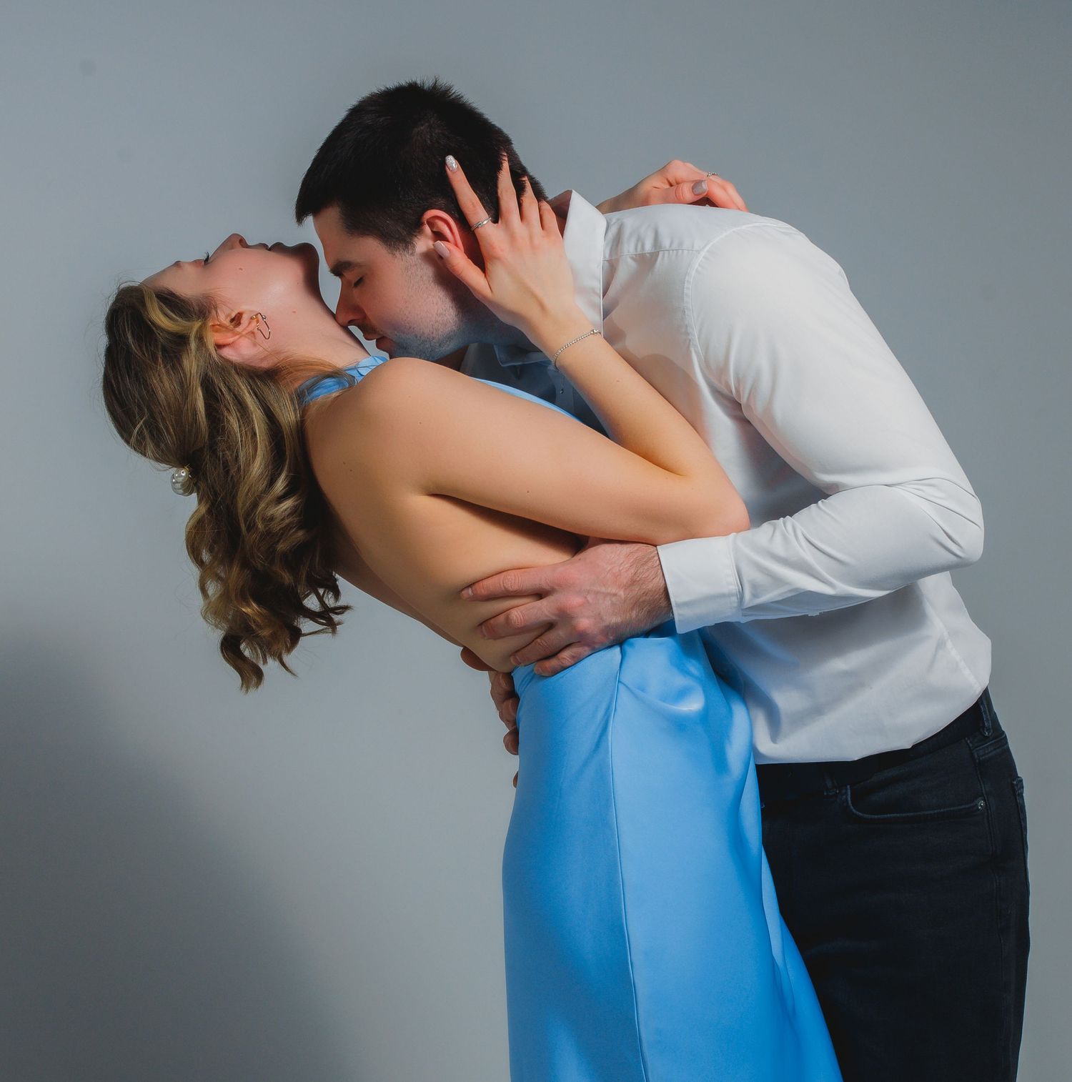 A man in a white shirt kisses a woman in a blue dress as she leans back in his arms against a gray background.