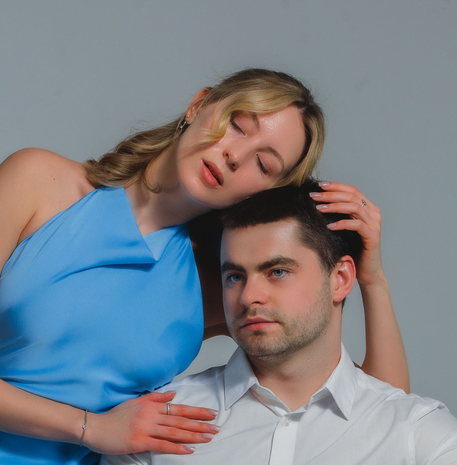 A woman in a blue top rests her head on the head of a man in a white shirt in a close, tender studio portrait.