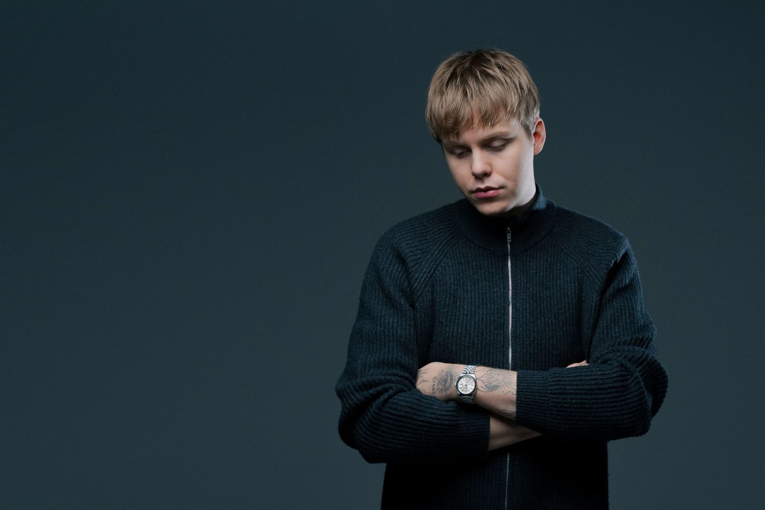 Portrait of a young man in a dark zip-up jacket with arms crossed, looking downward against a dark background