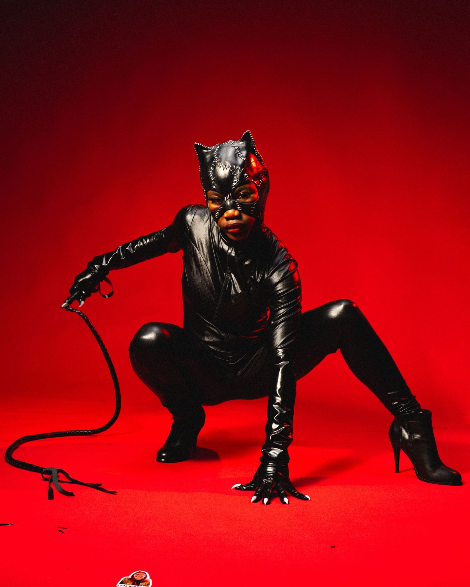Person in black latex Catwoman costume with whip, cat mask, and heels posing against a red background.