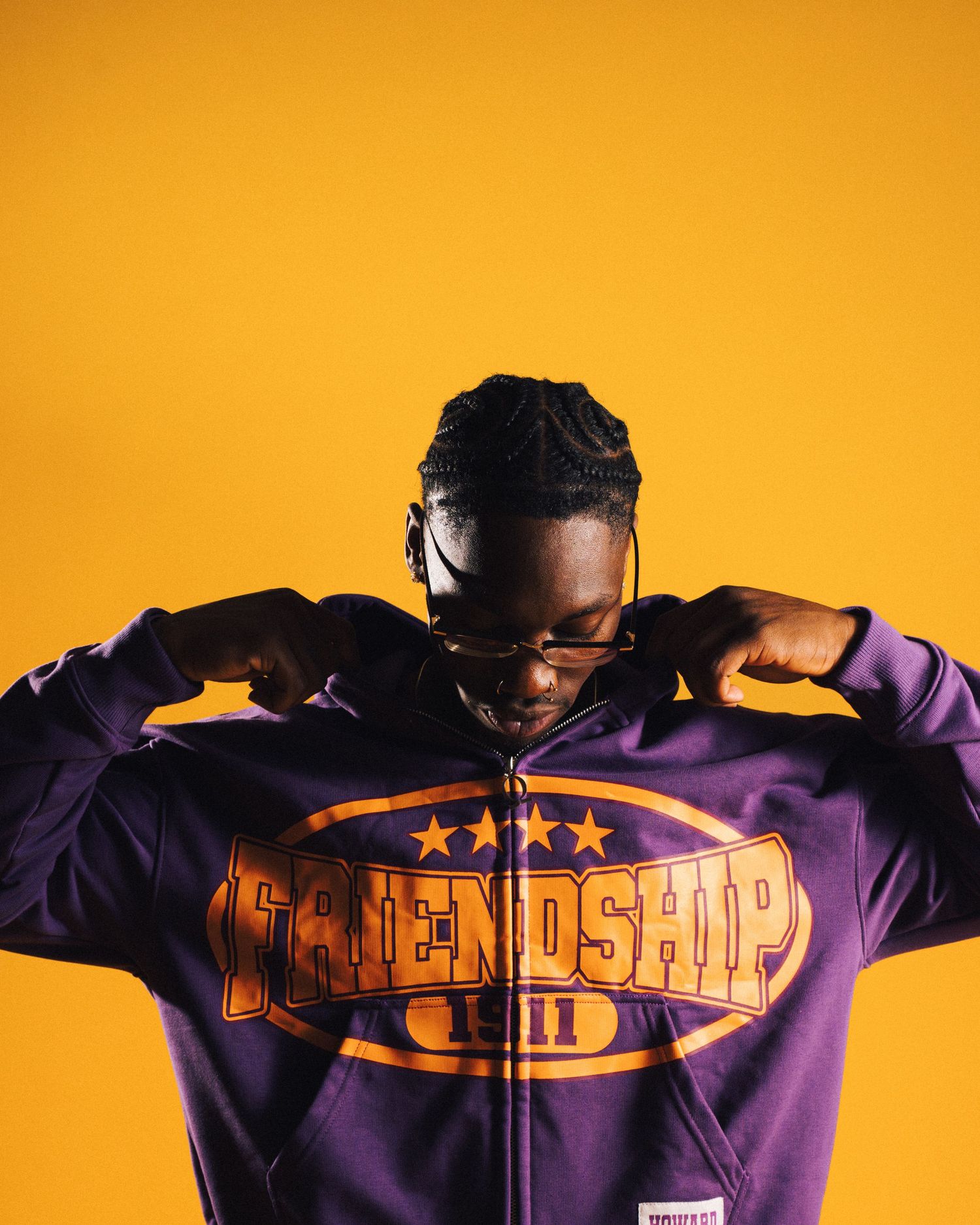 Man in purple Friendship 1911 hoodie posing against vibrant yellow background with braided hair and glasses.