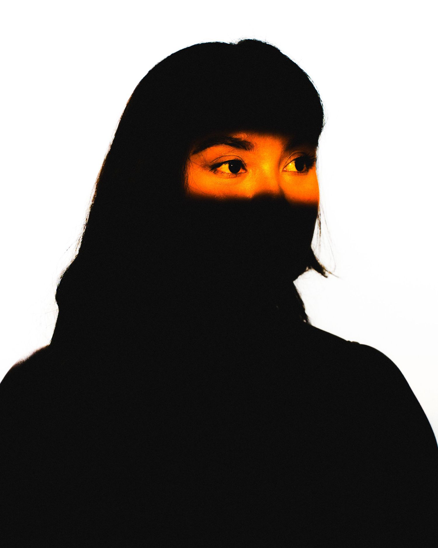 Dramatic silhouette portrait of a person with long dark hair, illuminated by warm orange light revealing their eyes.