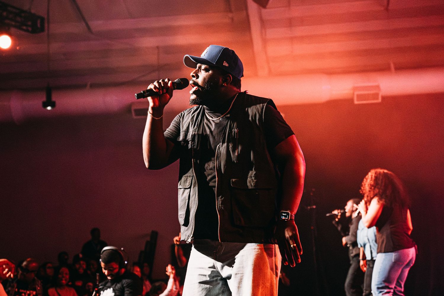 Male rapper performing on stage with microphone under red lights at a live music concert event.