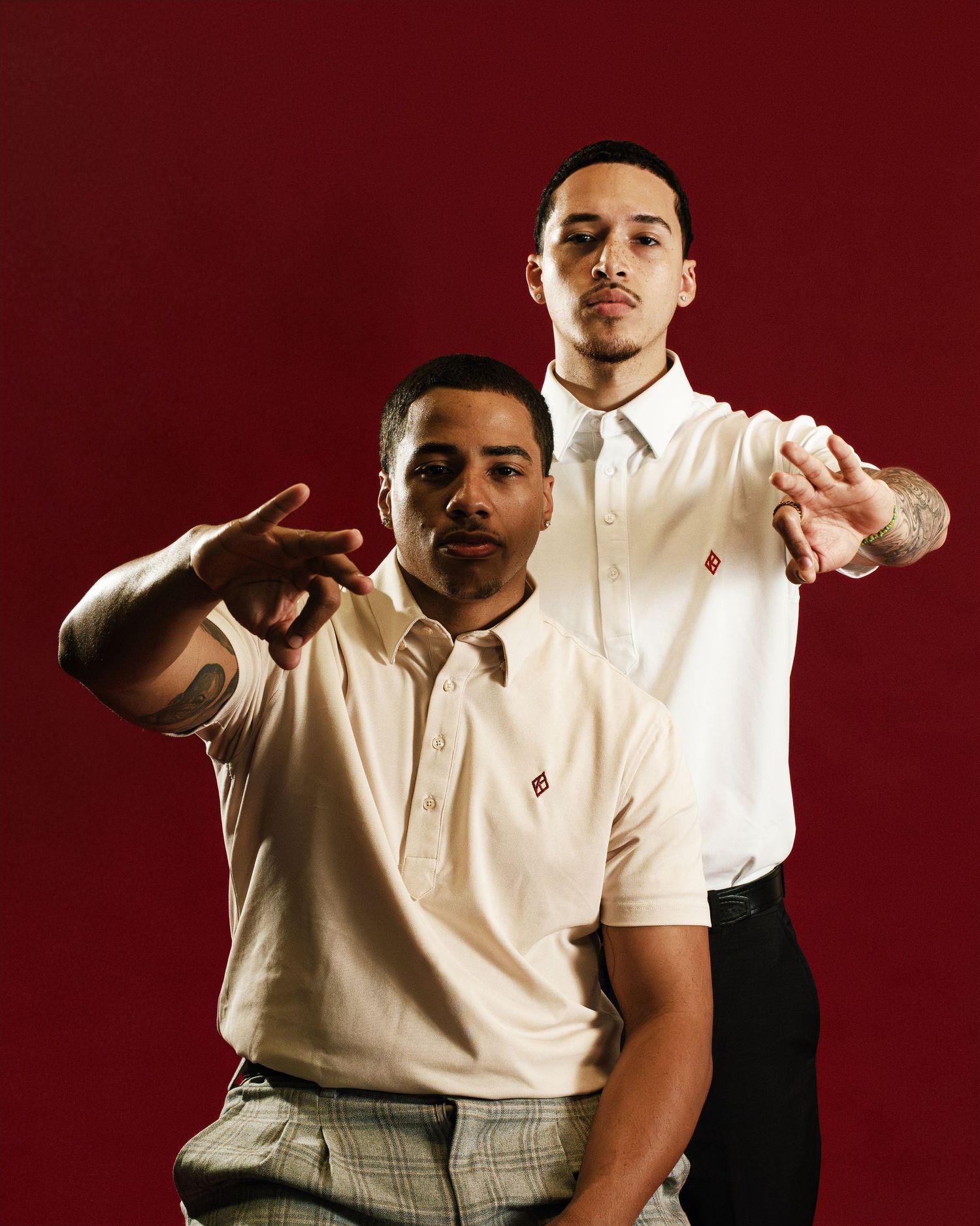 Two young men in cream shirts pose against a red background, pointing toward the camera with confident expressions.
