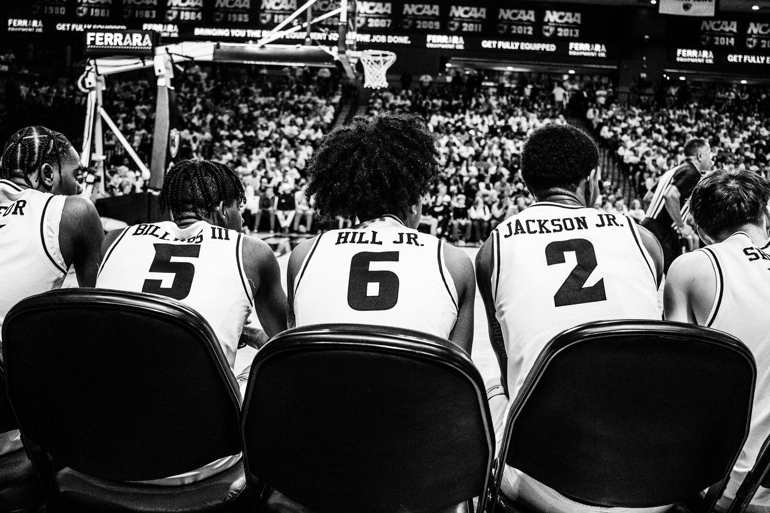 Three basketball players sit on chairs courtside wearing jerseys numbered 5, 6, and 2.