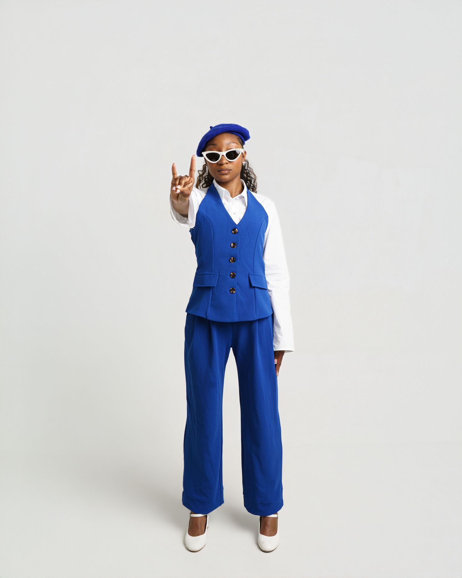 A woman in a blue vest, wide-leg pants, and beret poses with a peace sign and white sunglasses.