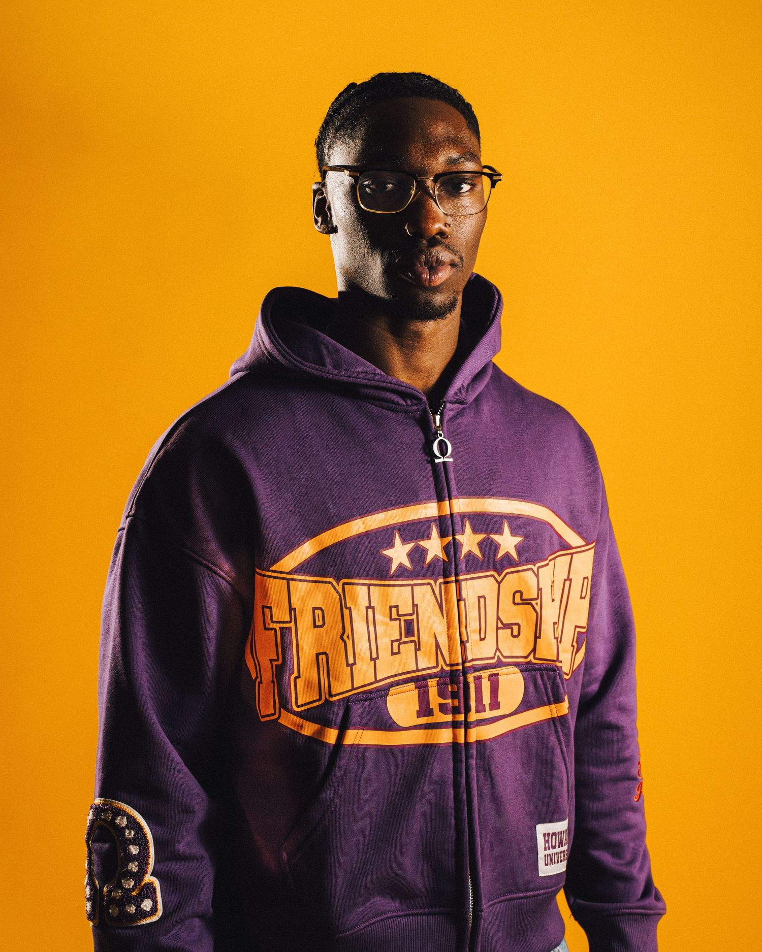 Man wearing purple Friends zip-up hoodie with glasses posed against bright yellow background.