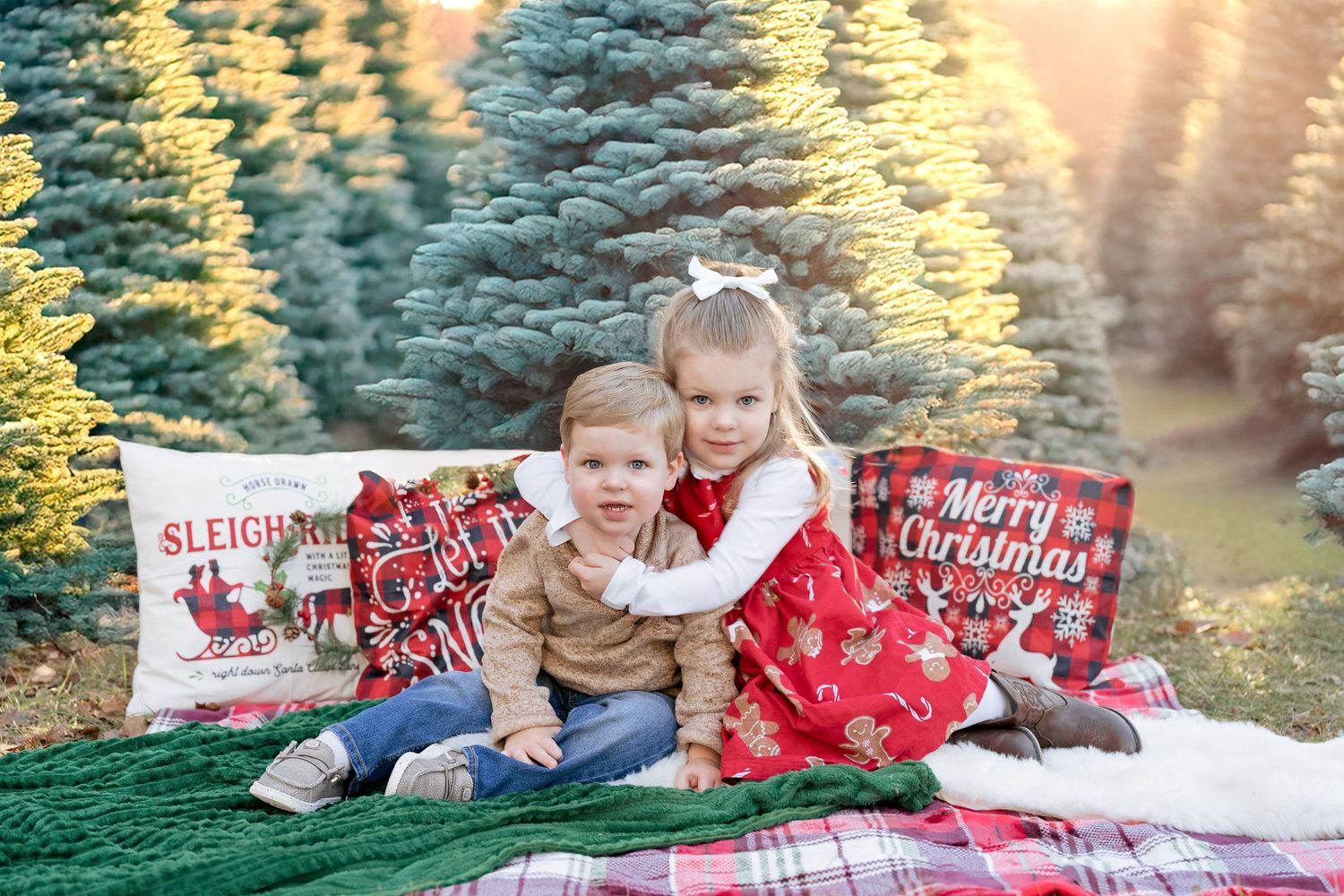 A young girl in a red dress hugs her younger brother while sitting on a festive blanket with Christmas-themed pillows