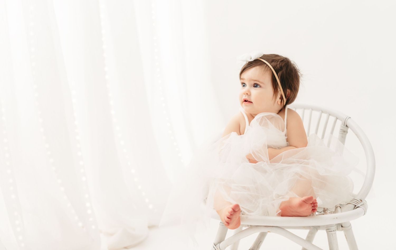 A small child in a white tutu dress sits thoughtfully in a vintage white chair against a light backdrop.