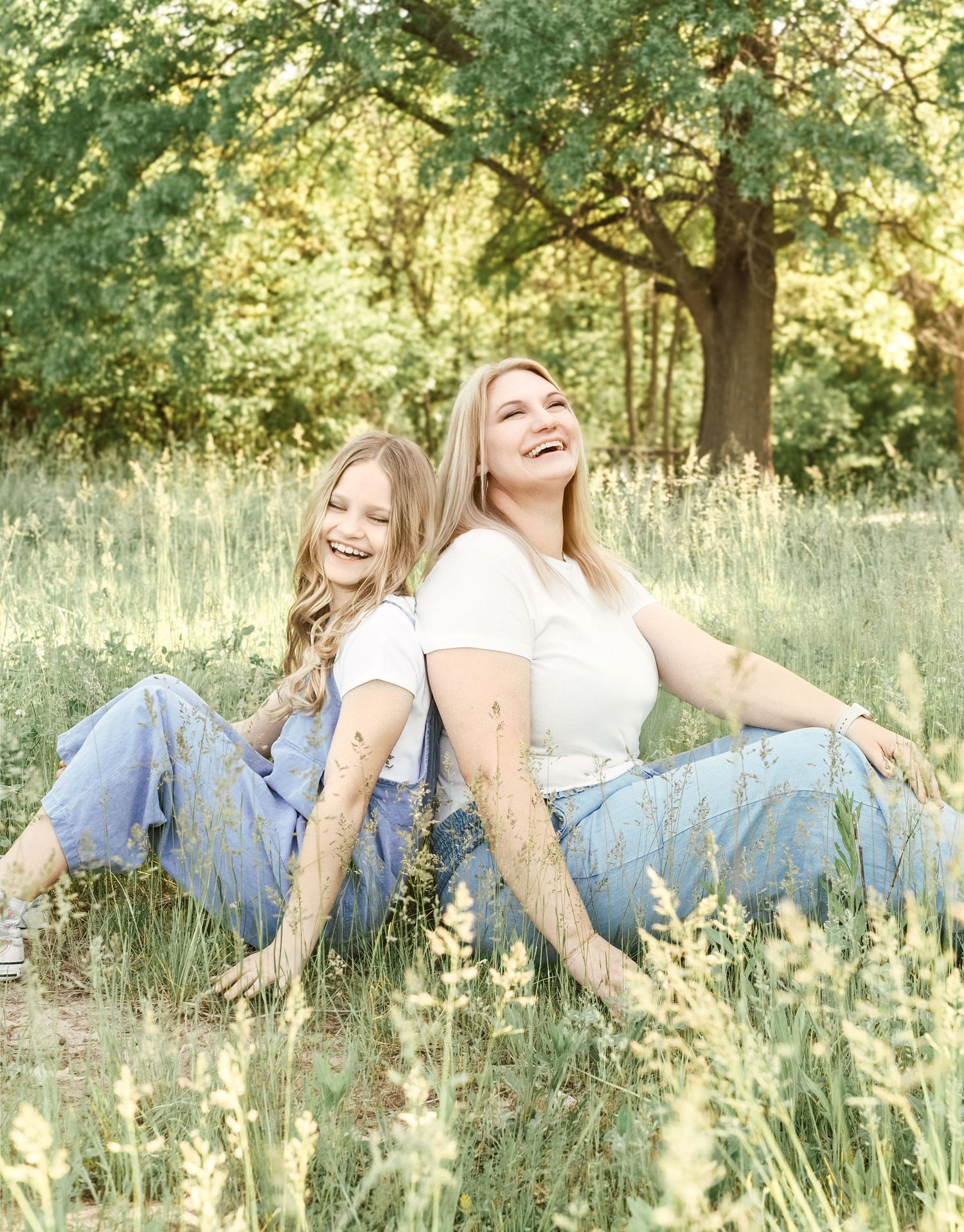 Two people in blue jeans and white tops sitting back-to-back laughing together in a sunlit summer meadow with tall grass.