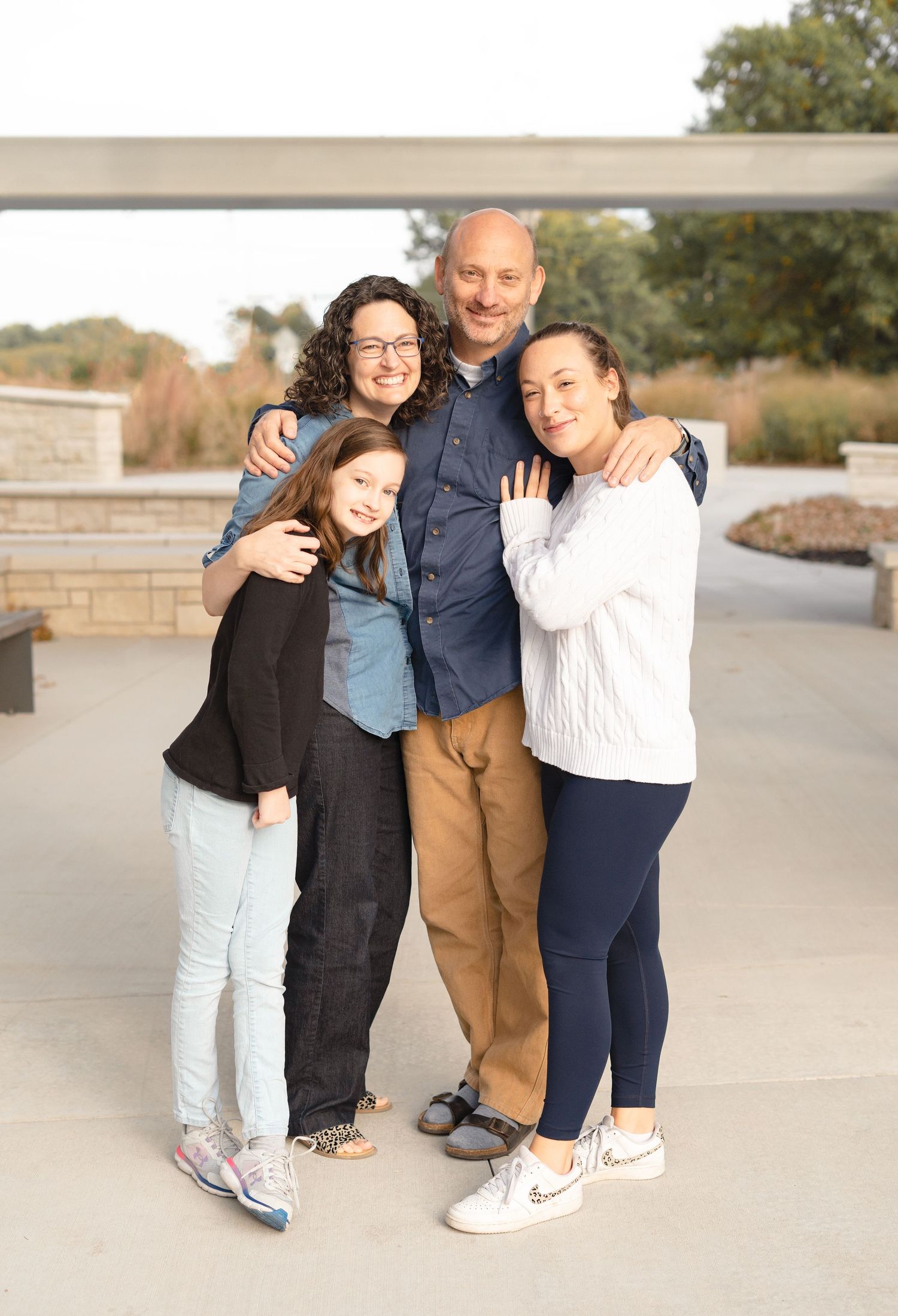 A family of four poses together outdoors on a sunny day wearing casual clothing and smiling at the camera.