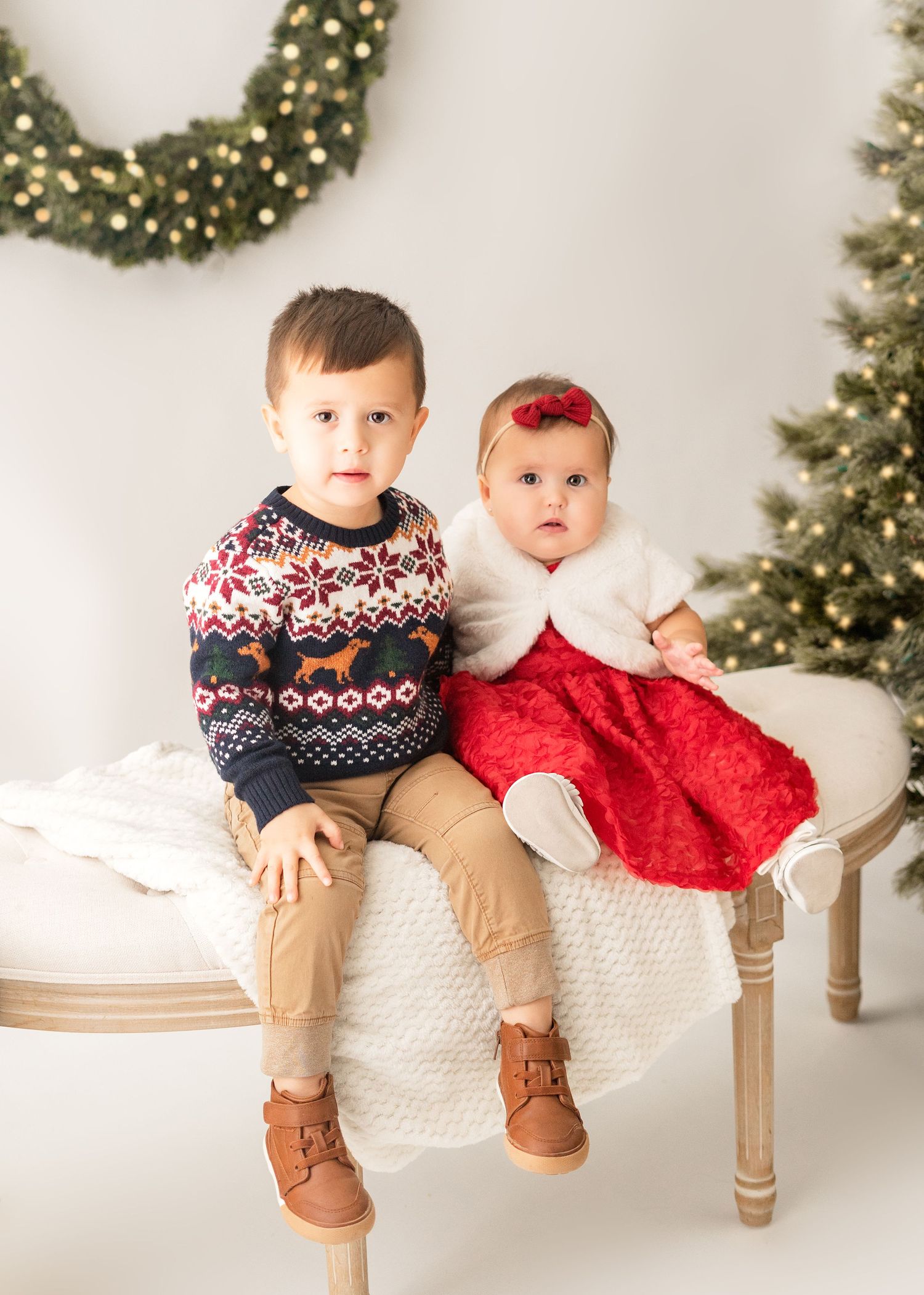 Two children in holiday attire sit on a white bench with Christmas decor and trees in the background.