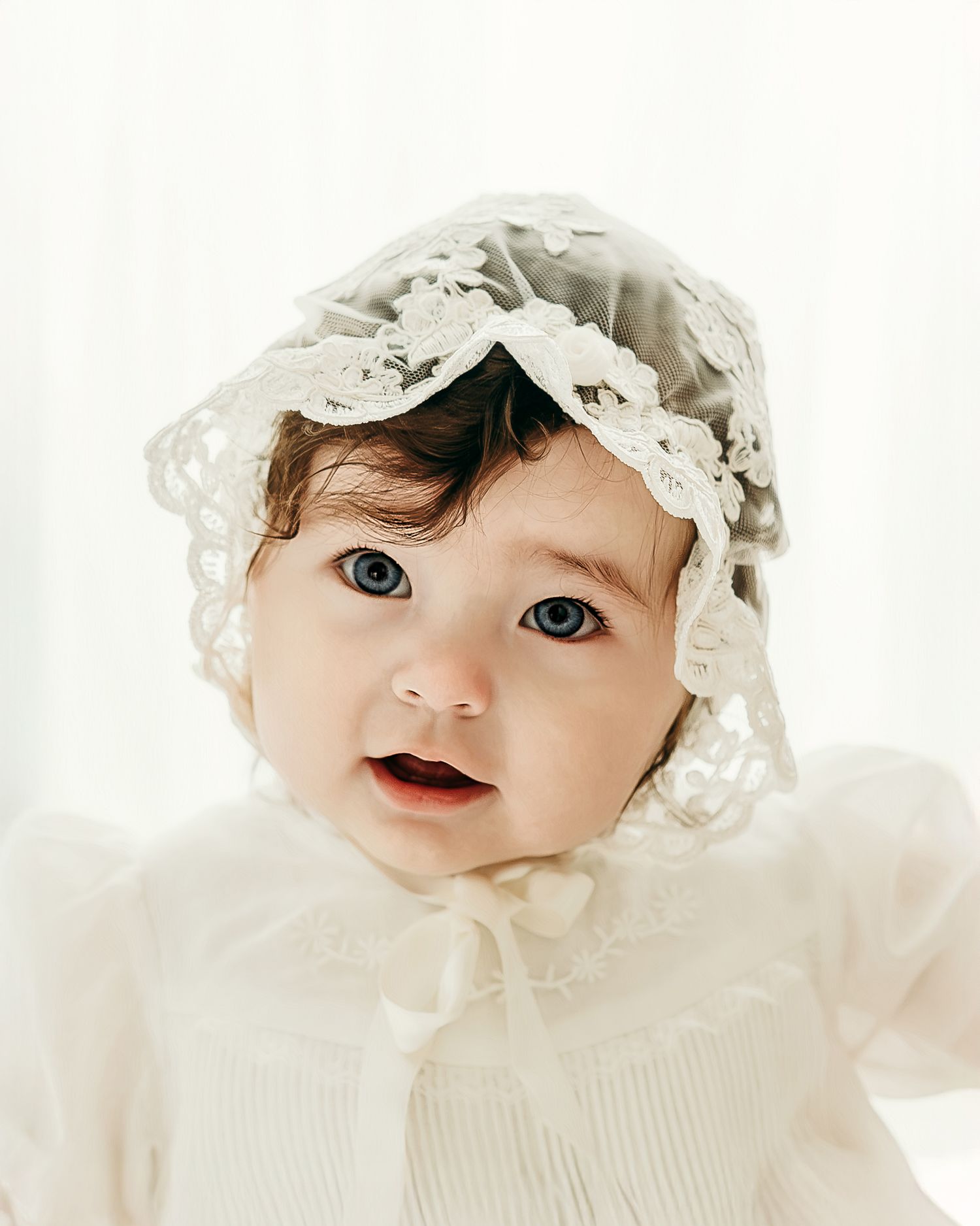 A baby girl wearing a lace bonnet looking toward the camera during a soft studio milestone session.