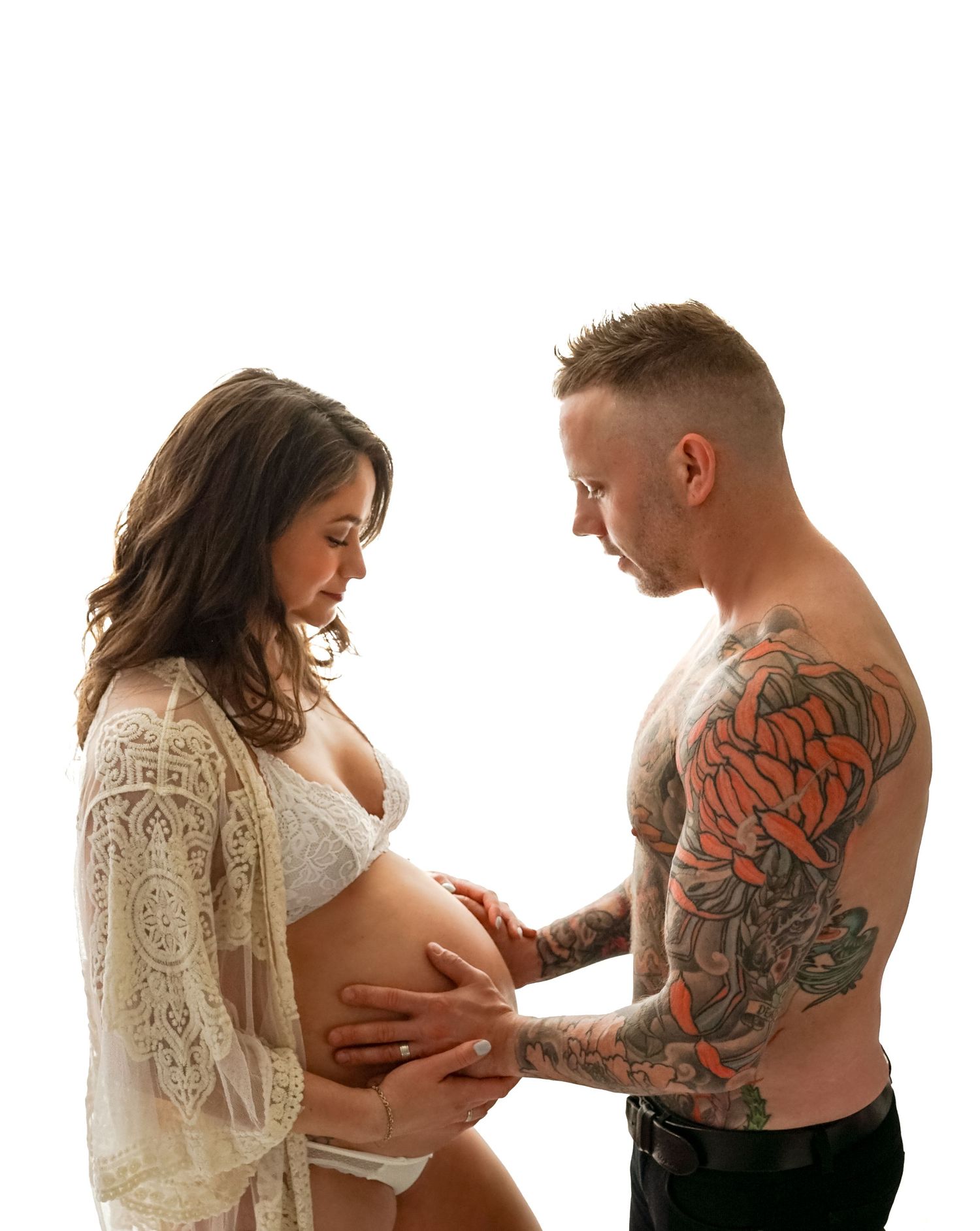 Pregnant woman in a white lace robe and partner with chest tattoo touching her bare baby bump in maternity photo shoot.