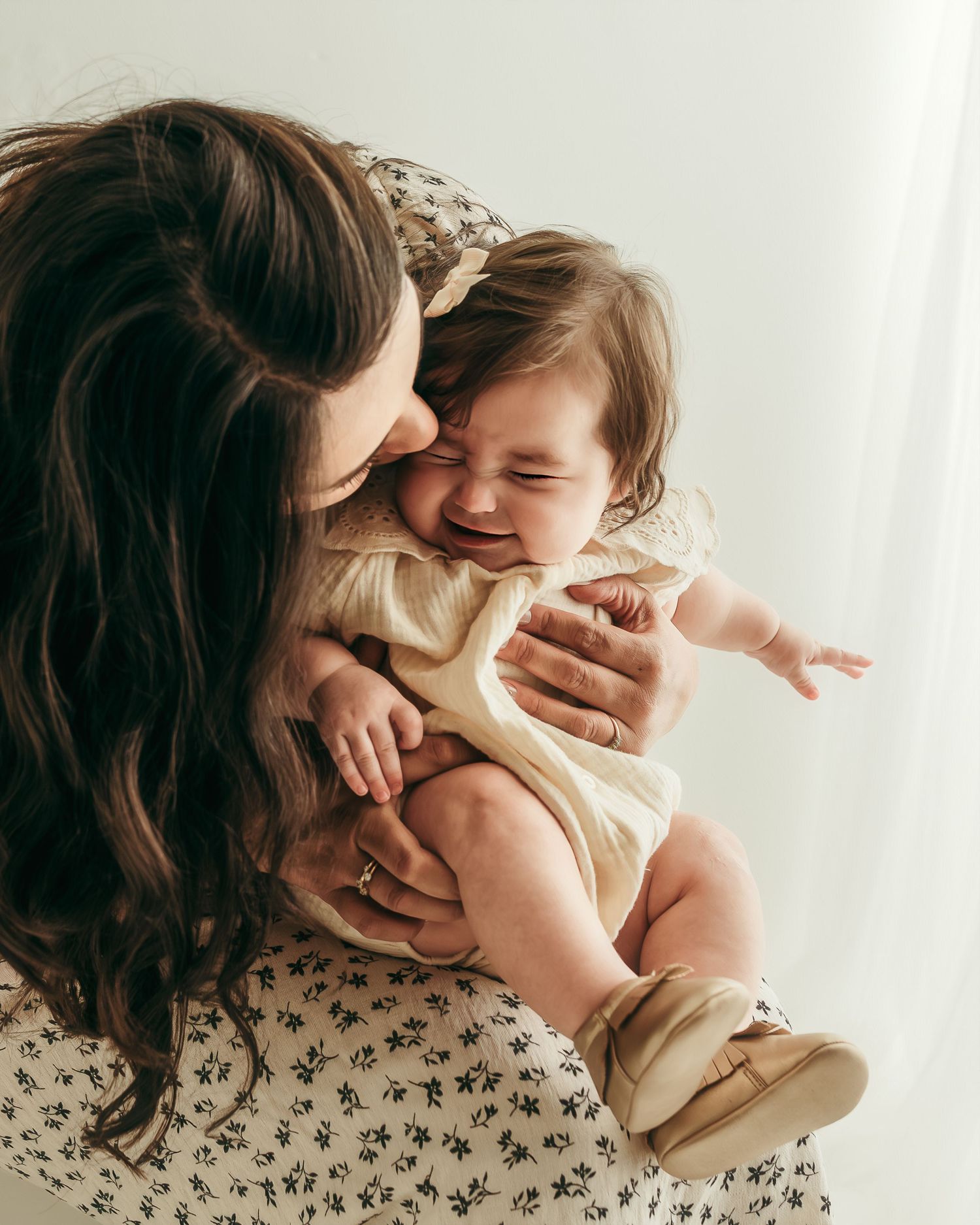 A joyful baby laughing while being held by a dark-haired mother in a floral dress against a soft white background.