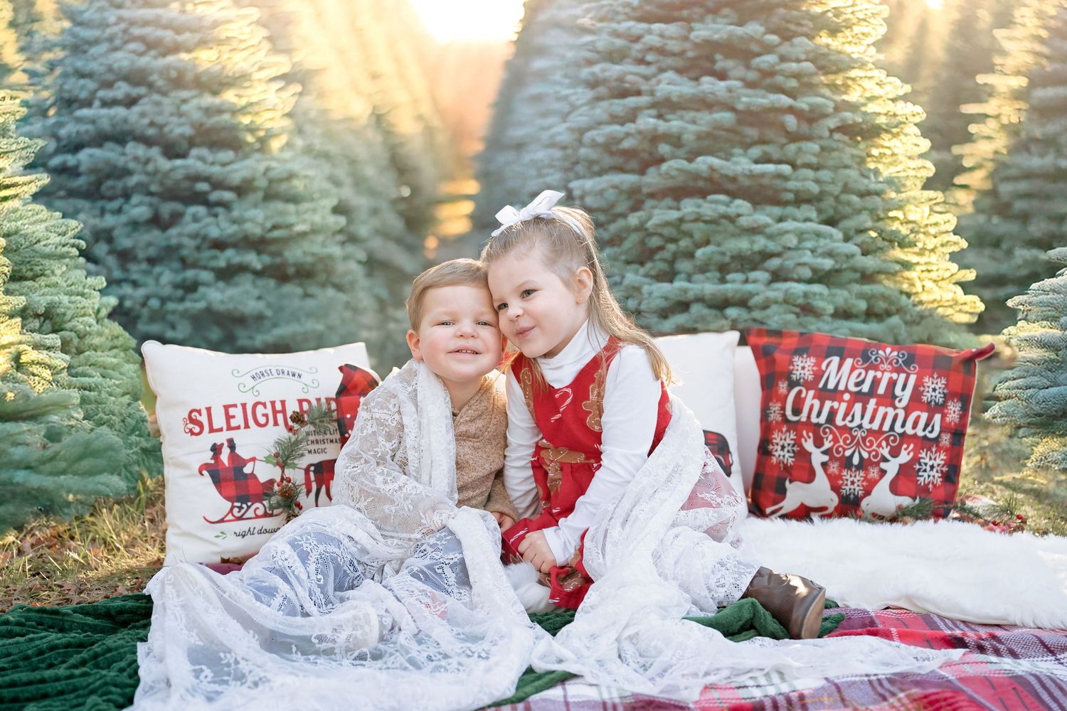 A young girl in a red dress hugs her younger brother while sitting on a festive blanket with Christmas-themed pillows