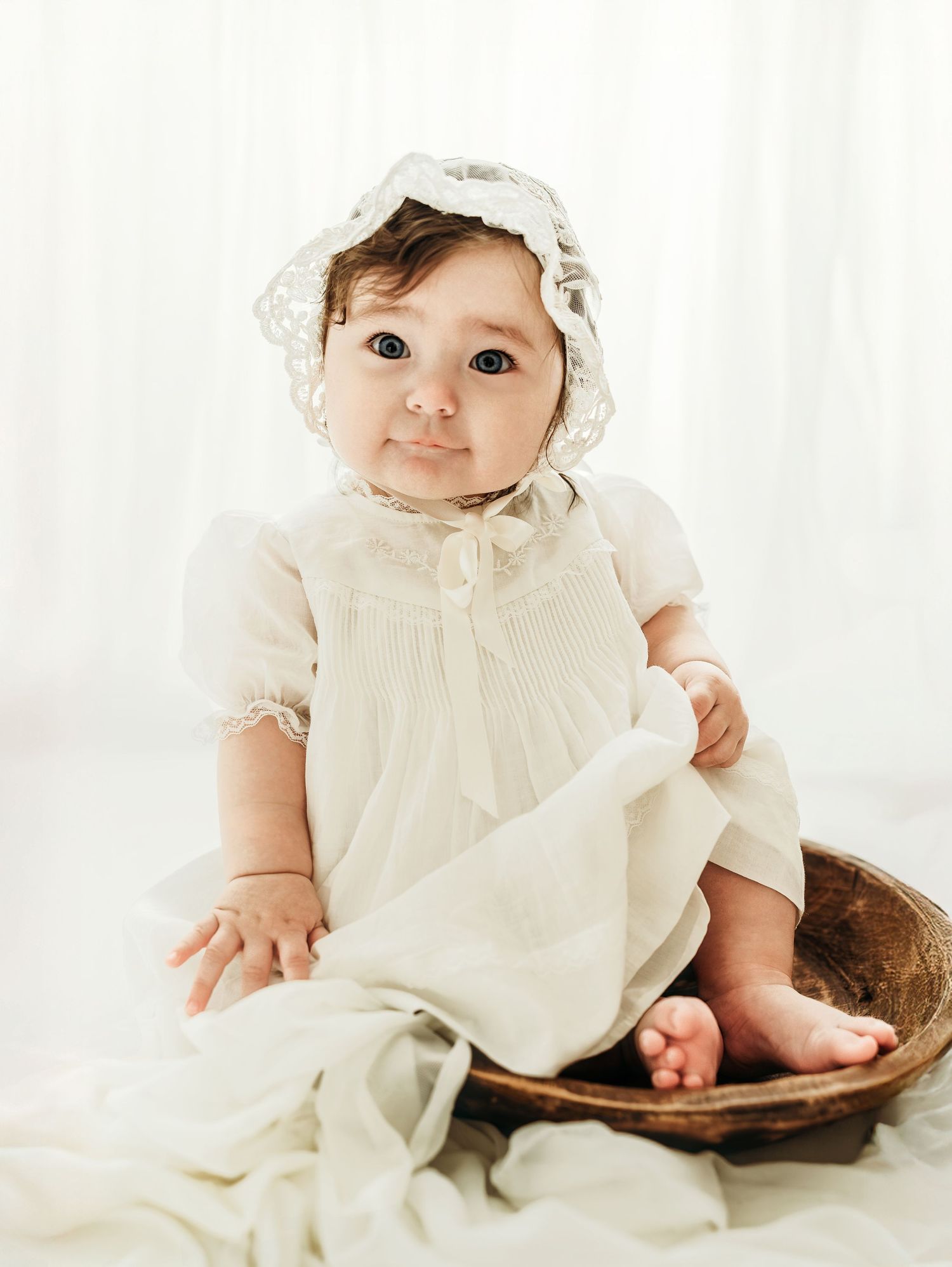 A baby girl sitting on a soft blanket wearing a cream dress during a studio milestone session.