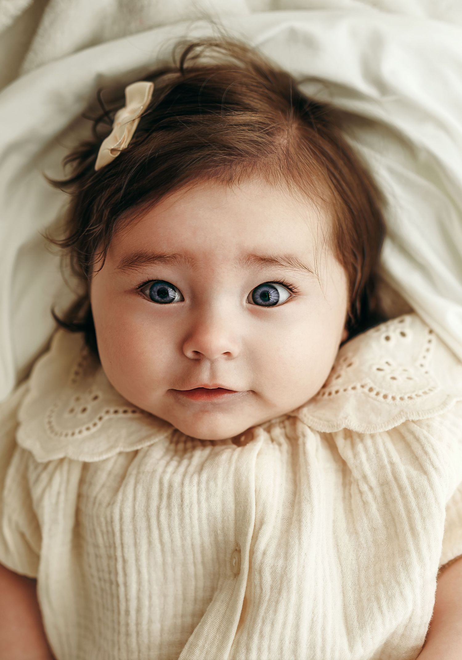 A 6 month old baby girl sitting and smiling in a neutral studio setting, wearing a soft cream outfit during a milestone photo session.