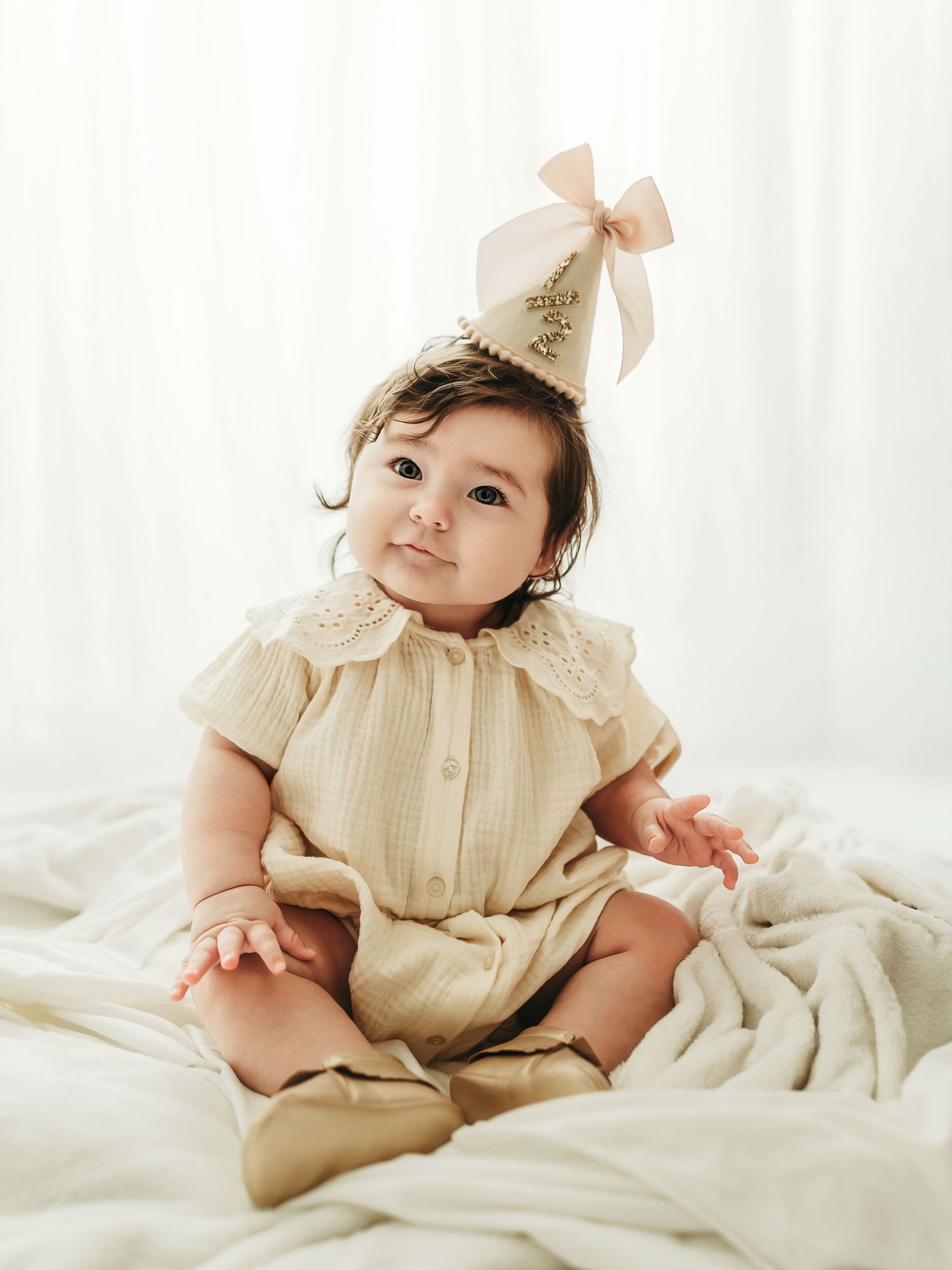 A smiling 6 month old baby girl sitting on a white backdrop during a soft, neutral milestone photo session.