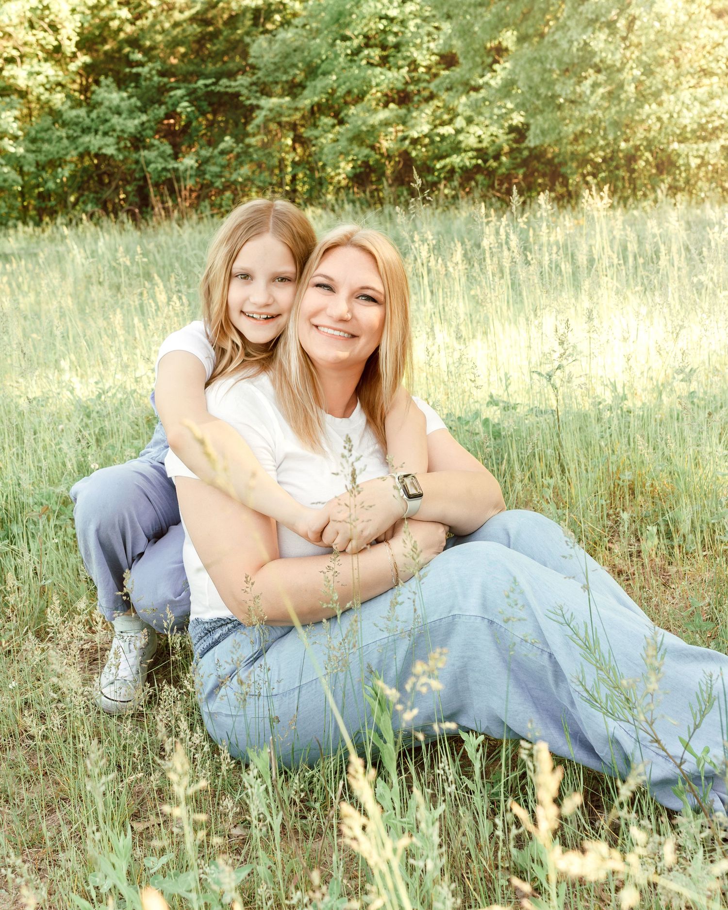 Two people sitting together and embracing in a sunny meadow with tall grass and trees in the background.