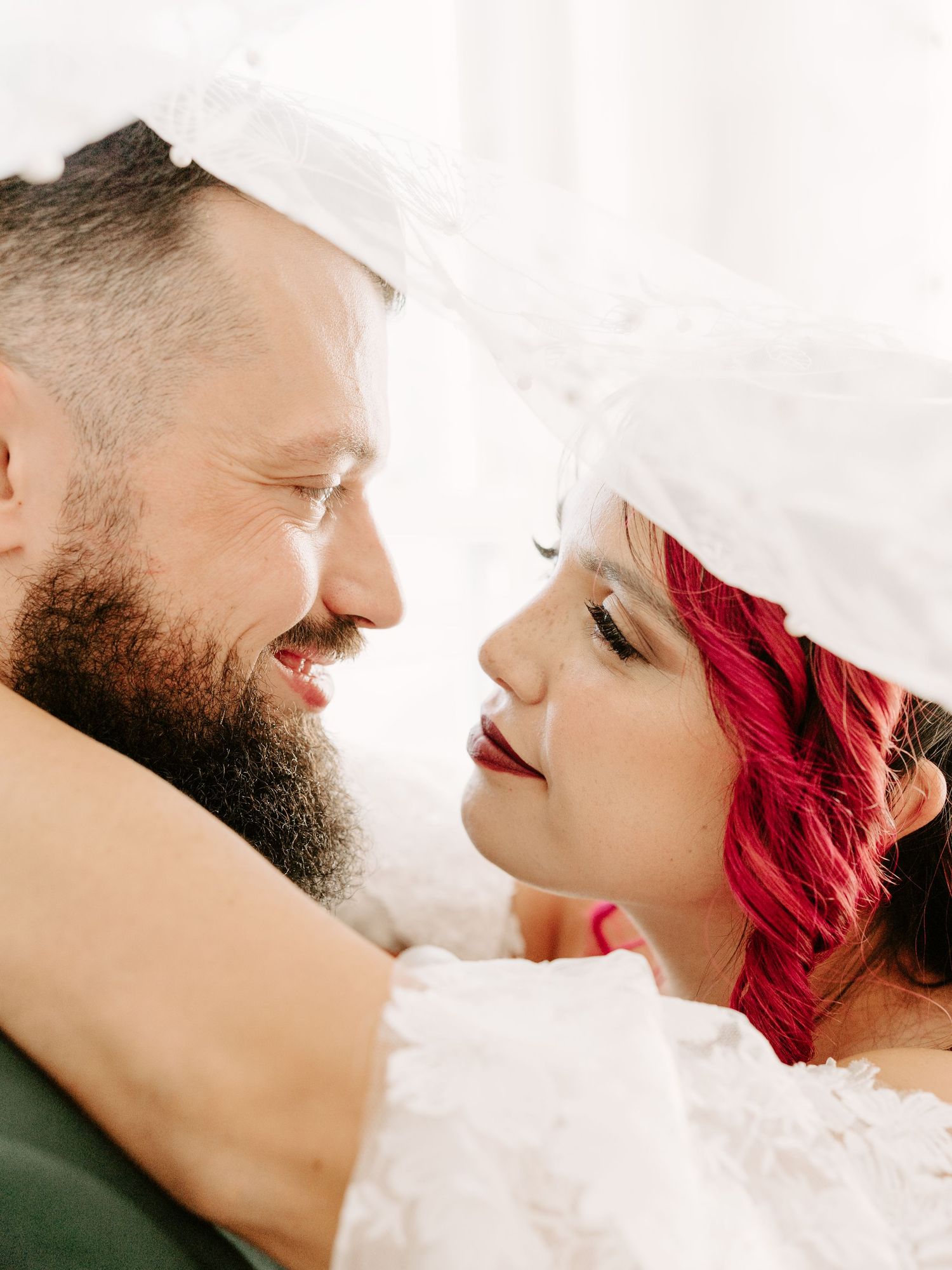 Romantic wedding photo under white veil with bride's vibrant red hair and groom's full beard as they share an intimate moment.