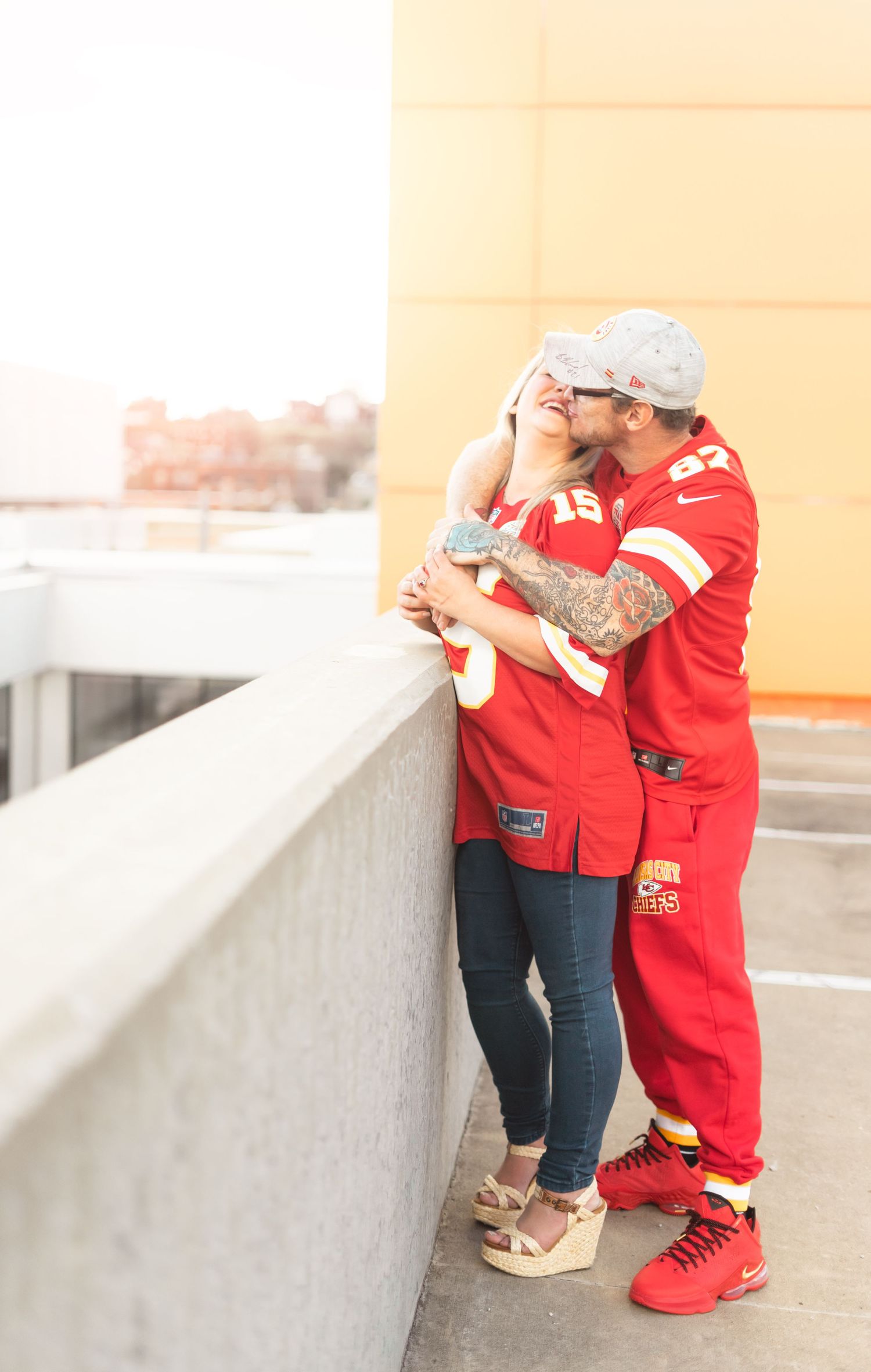 A couple in KC chiefs jersey's share a tender moment on a rooftop balcony during a sunny afternoon.