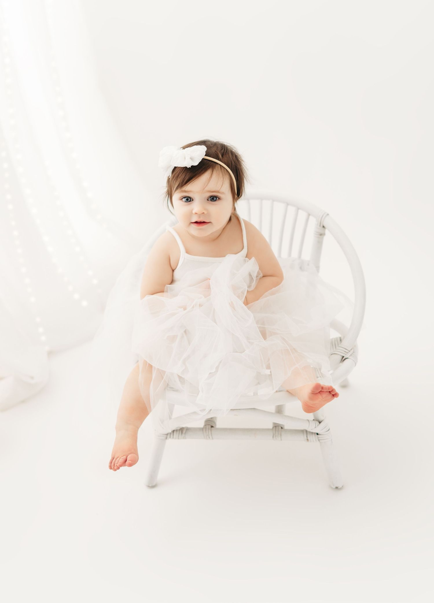A small child in a white tulle dress sits on a white wicker chair against a light background.