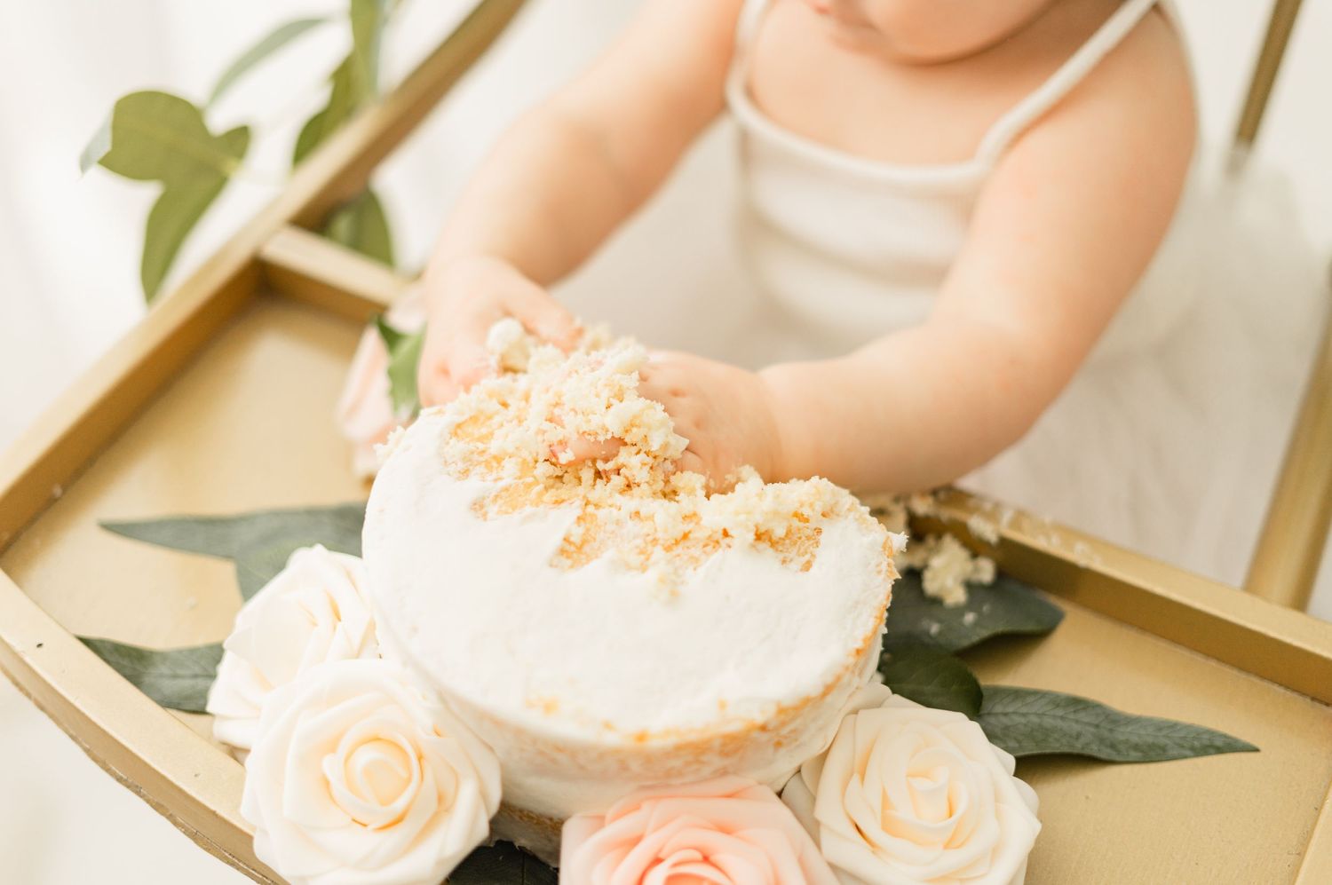 A white frosted cake decorated with crumbs sits on a table with delicate cream-colored roses and greenery.