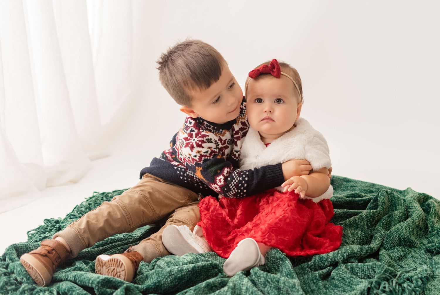 Two children in holiday outfits sit together on a green velvet blanket against a white backdrop.