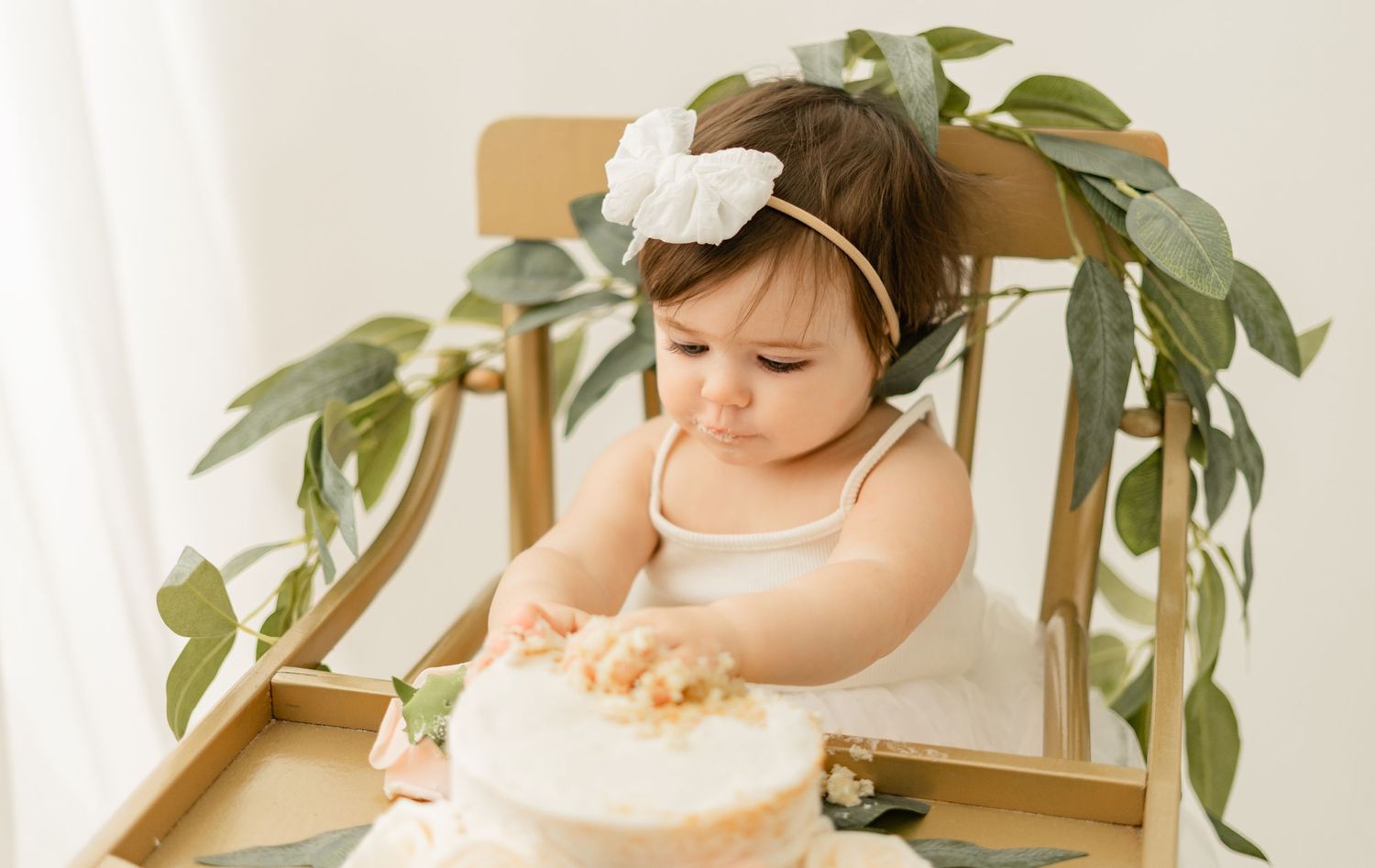 A small child in a white top sits in a wooden high chair decorated with green foliage, reaching for a cake.