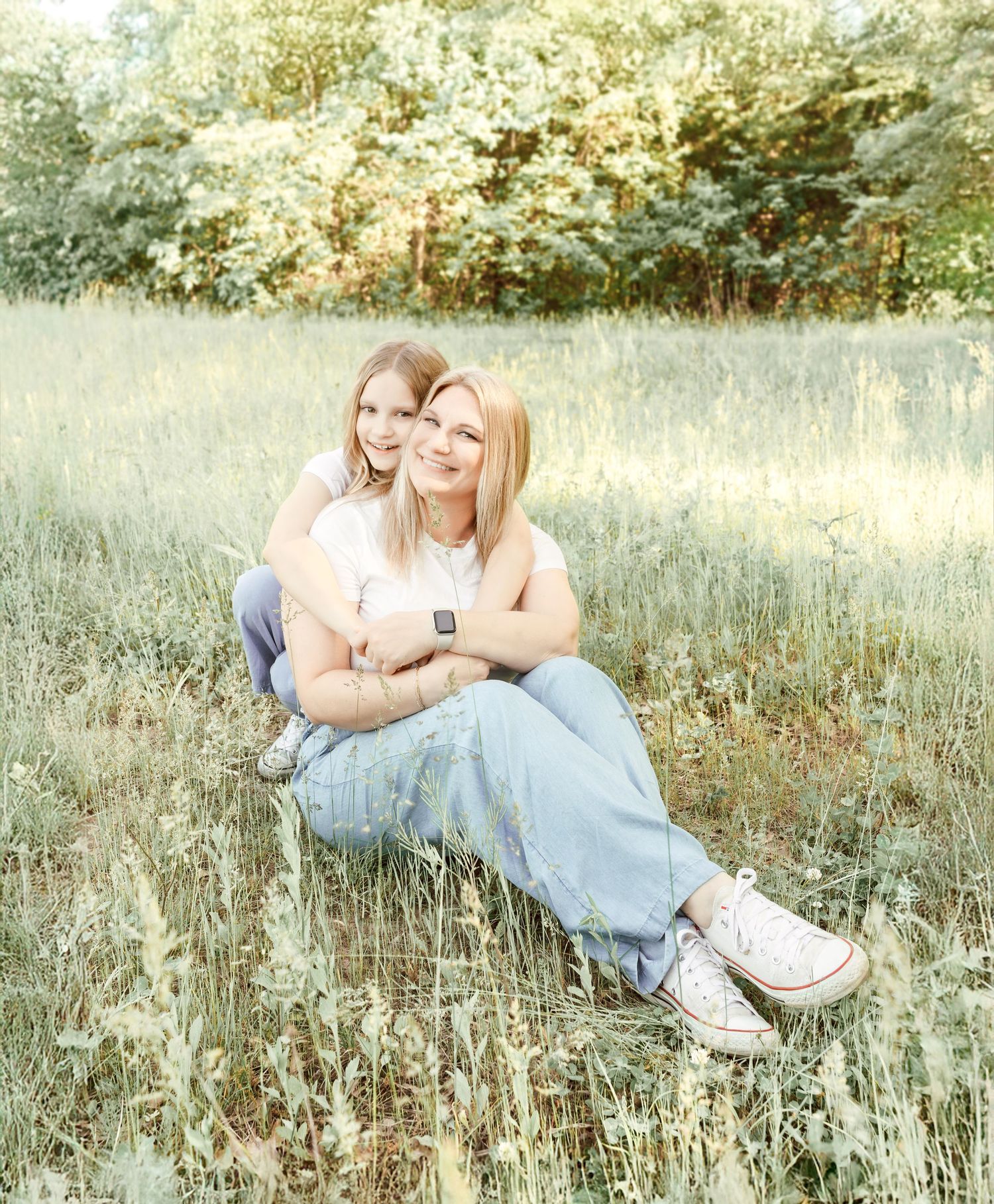 Two people sit close together in a dreamy meadow with tall grass and trees during golden hour.