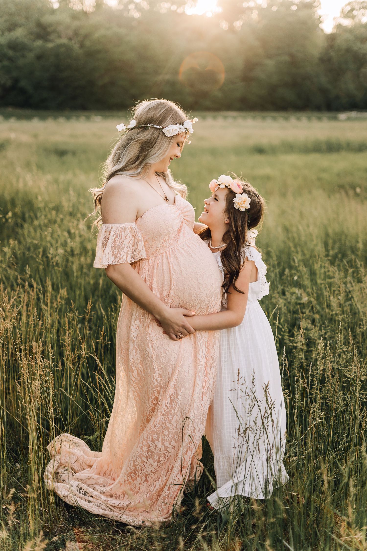 A pregnant woman in pink lace gown and child in white dress share tender moment in sunlit meadow at sunset.